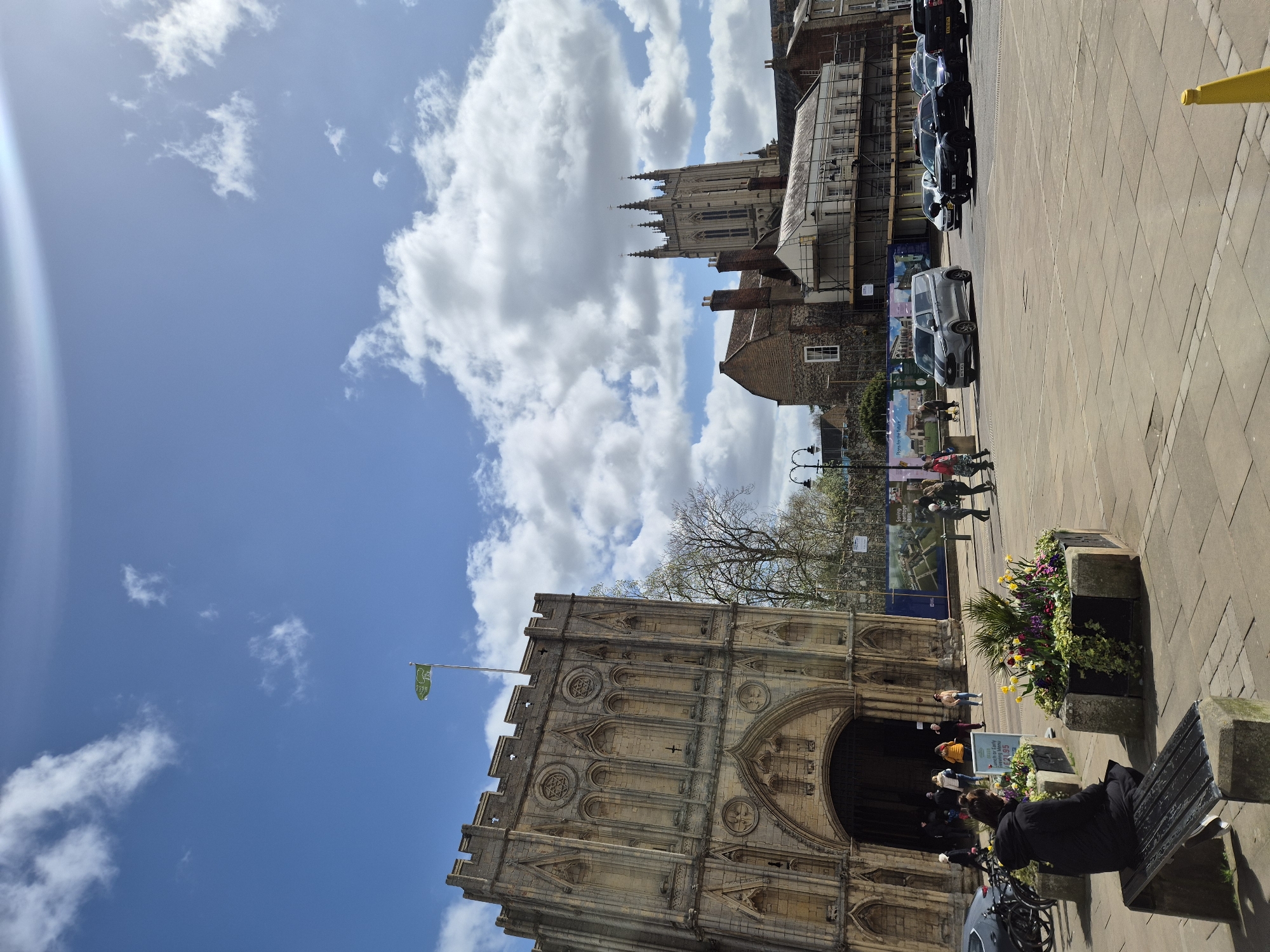 The image shows the Angel Hill area in Bury St Edmunds, West Suffolk, United Kingdom. The main subject is the iconic Abbey Gate, a large stone archway with intricate carvings. A green flag flutters atop the gate. People are walking and gathering near the entrance, suggesting pedestrian traffic. To the right of the gate, there's a stone bench with a person sitting on it. Flowerbeds add color to the scene. Behind the Abbey Gate, to the left, the Bury St Edmunds Cathedral with its gothic architecture is visible in the background, partially obscured by the sky with scattered clouds. On the right side of the frame, a row of parked cars lines the paved street. Construction or renovation work is evident from the presence of scaffolding and partially obscured buildings near the cathedral.