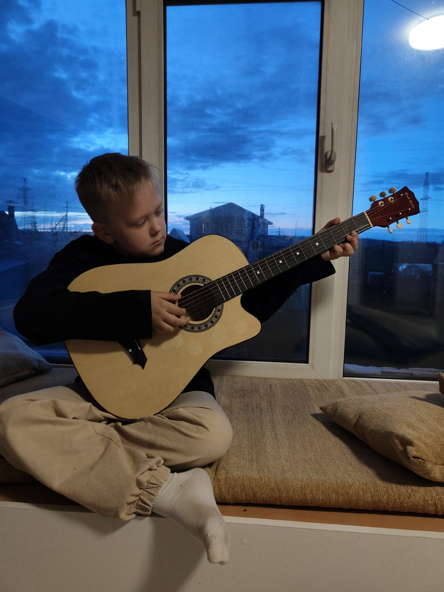 A boy sits cross-legged on a cushion in front of a large window, playing an acoustic guitar. He wears a black long-sleeved shirt, beige pants, and one white sock. Outside the window, a twilight sky is visible above buildings and power lines. A pillow rests to his right on the cushion.