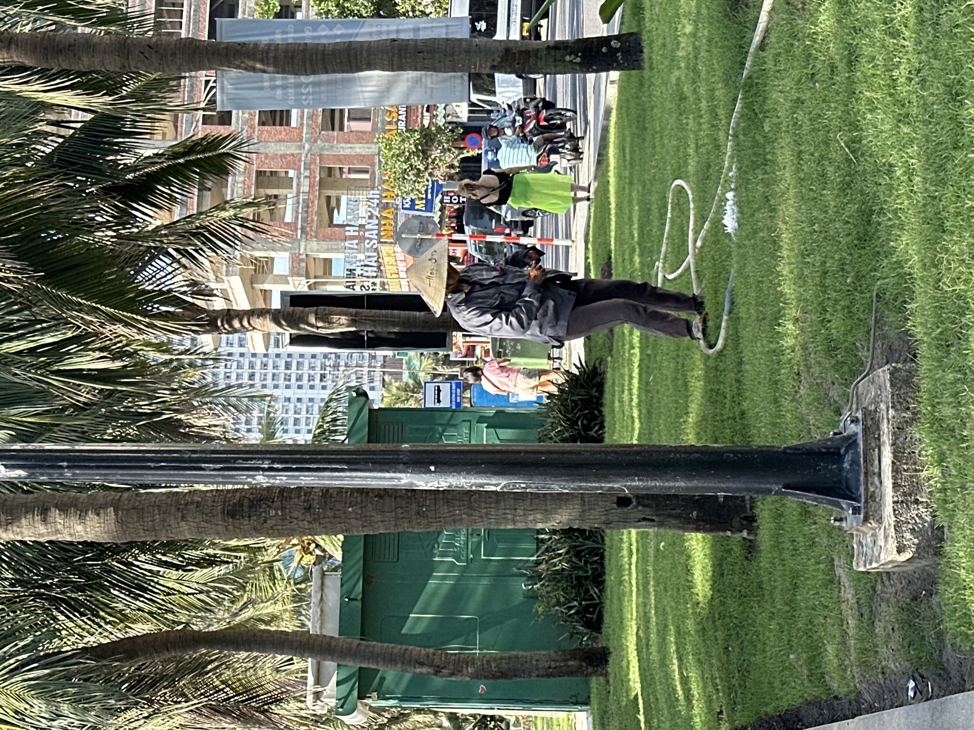 The image captures a street scene in Đà Nẵng, Vietnam, with palm trees lining a grassy area. In the foreground, a person wearing a conical hat and a dark jacket is tending to a white hose lying on the grass, with some water splashing. Behind them, a woman in a bright green skirt walks away from the camera, followed by a man on a motorbike. To the left, a green utility structure with visible text and a bus stop sign are present. Construction sites and tall buildings are visible in the background, indicating an urban environment. The overall atmosphere is sunny and appears to be during daylight hours.