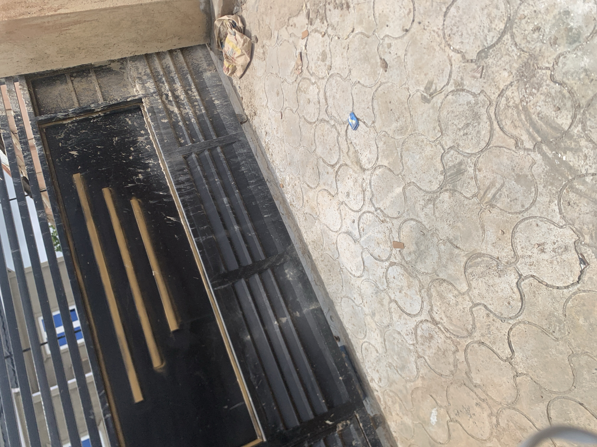 The image depicts a street-level view of a black metal gate, possibly leading into a property, with horizontal louvers. Three yellow-gold horizontal bars are inset into the upper section of the gate. The gate appears weathered and somewhat dirty. To the right of the gate, a beige-colored concrete pillar stands, also showing signs of wear and dust. The ground in front of the gate is paved with interlocking concrete blocks, exhibiting a patterned arrangement. A crumpled brown paper bag, possibly containing construction materials, lies on the paving stones near the pillar. A small, blue object, possibly a wrapper or packaging, is visible on the paving stones further away from the gate. Debris, including small pieces of what appears to be brick or tile, is scattered across the ground. In the background, through the upper louvers of the gate, glimpses of a building with blue window frames and a roof are visible, suggesting an urban or semi-urban setting. The lighting suggests it is daytime, with shadows indicating the sun's position. The overall impression is of an entrance to a residential or commercial property in an area that may be undergoing construction or maintenance. The scene is consistent with locations found in the city of Jos, Nigeria, given the architectural style and material common in the region.