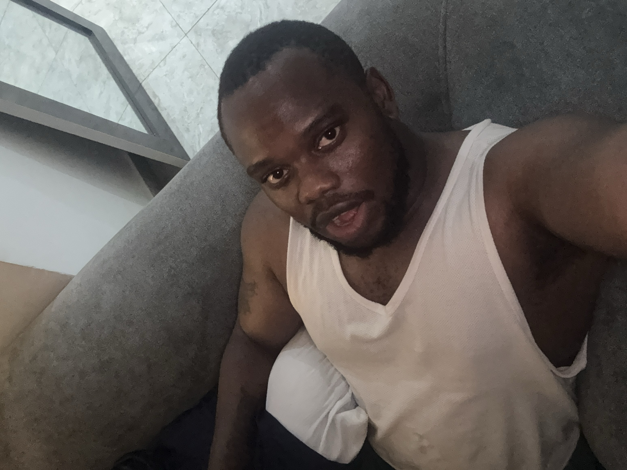A man is lying on a gray couch, wearing a white tank top. He has short, dark hair and a beard. The background includes a floor with gray tile and a metal frame. The location is unknown and could not be determined.