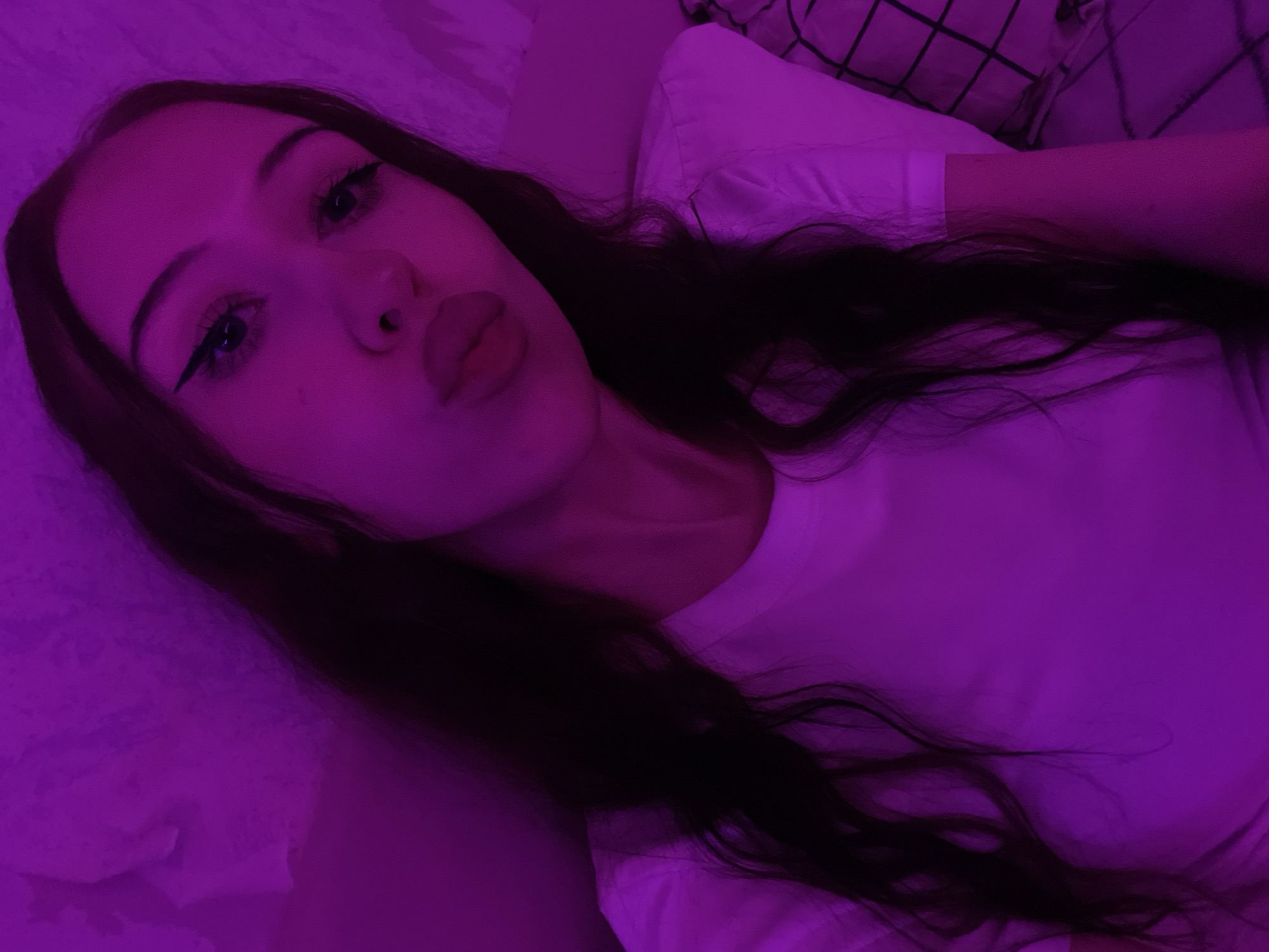 The media file shows a close-up, overhead shot of a young woman lying down, illuminated by purple lighting. She has long, dark hair that is spread out around her. Her face is the focal point, with her eyes looking towards the camera and her lips slightly pursed. She is wearing a white t-shirt, and the background appears to be a bed with pillows. The overall mood is intimate and relaxed, possibly taken at night or in a dimly lit room. There is no visible text or weather cues. The location is Votkinsk, Russia.