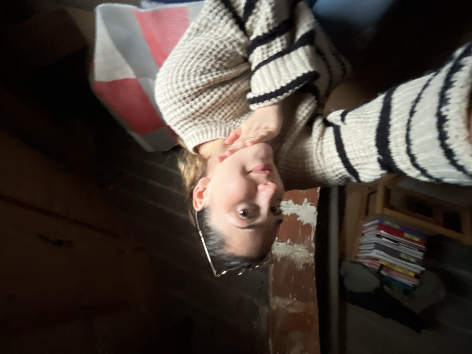 The image depicts an inward-facing camera perspective of a young adult female located indoors, within a structure in Ancenis, France. The subject, positioned off-center and angled slightly, looks directly at the camera with a subtle smile. She has dark hair pulled back, with eyeglasses resting on her head, and wears a light-colored, waffle-knit sweater with black horizontal stripes on the sleeves. Her right hand is raised to her jawline, exhibiting a ring on one finger, while her left arm extends towards the camera. The immediate background above and to the left of the subject features dark, rough-hewn wooden planks or beams, indicative of an unfinished ceiling or wall. Below this, a section of lighter-colored, possibly painted brickwork is visible. To the far left, a stack of colorful books or magazines rests on a low, light-wood shelving unit, beside a dark, indistinct object. To the right of the subject, behind her shoulder, a multi-colored object, appearing to be a cushion or folded fabric with red, white, and grey sections, is partially visible. The lighting is dim, suggesting an enclosed or poorly lit interior space.