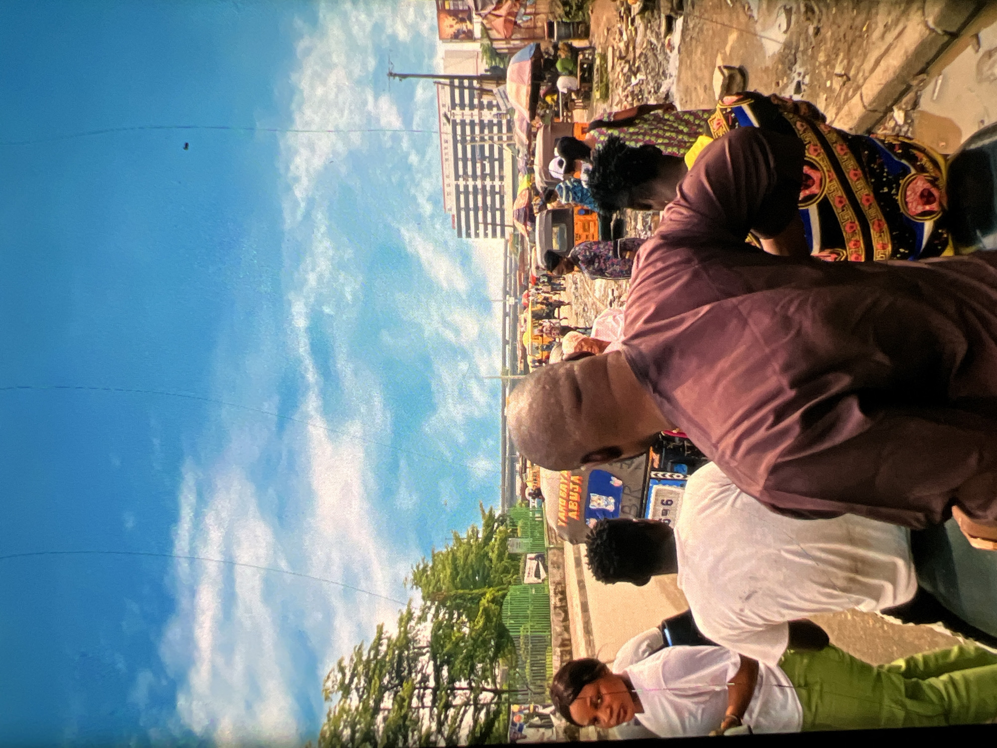 The image depicts a busy street scene in Lagos, Nigeria, under a bright blue sky with scattered clouds. In the foreground, several individuals are visible, some seated on what appears to be a motorcycle or tricycle. A woman in a white t-shirt and green pants is looking towards the viewer. Behind her, two men are seated facing away, one in a white t-shirt and another in a maroon traditional outfit. Further back, the street is filled with various modes of transportation, including yellow tricycles and other vehicles. Numerous people are walking and engaging in activities, with market stalls and umbrellas set up along the roadside. A multi-story building is visible in the background, along with trees and power lines. The ground appears wet and muddy in places, suggesting recent rain or a drainage issue. The overall impression is one of a vibrant and active urban environment.