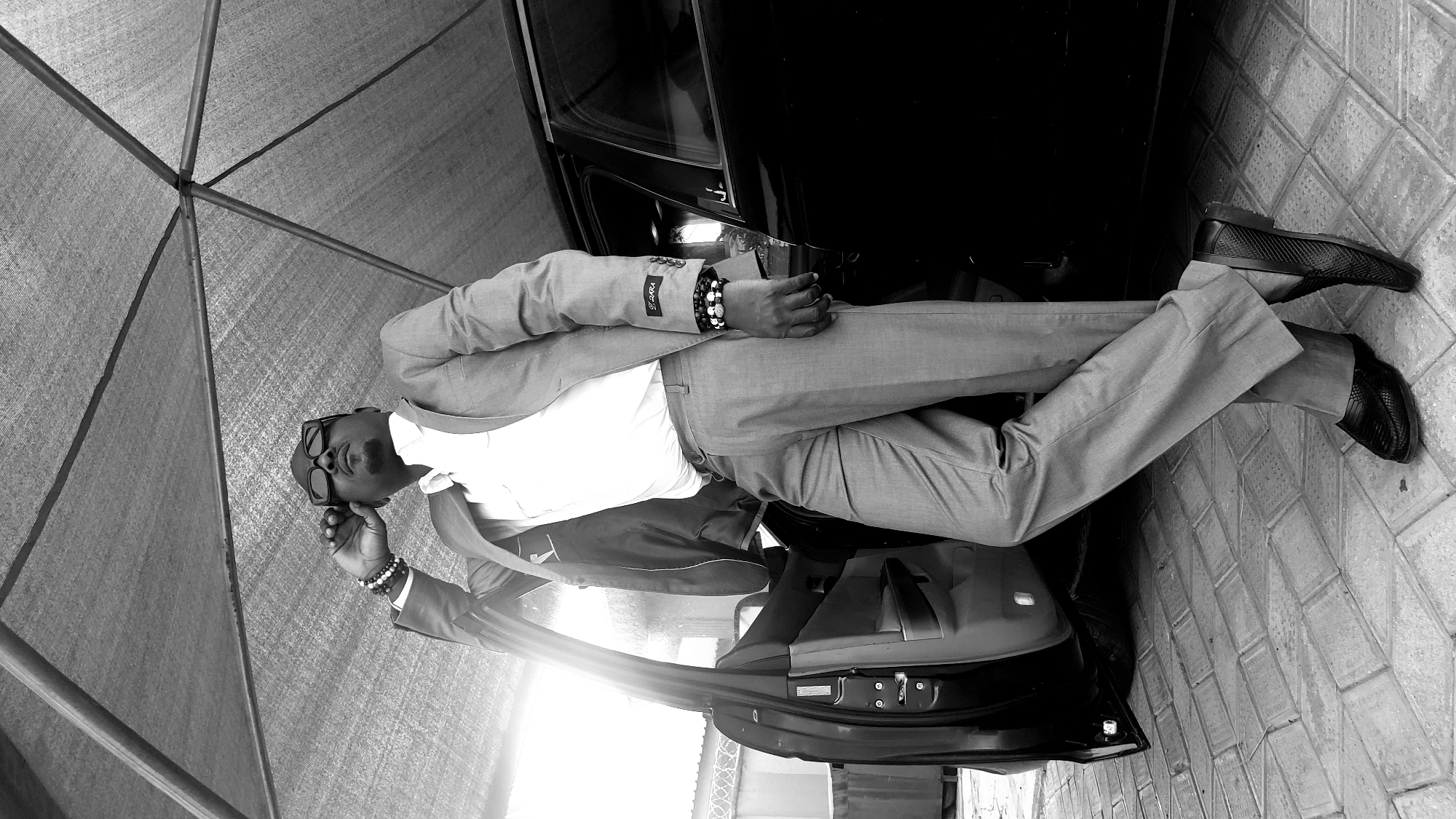 This black and white, portrait-oriented image captures an adult male seated by a black car, viewed from a slightly low angle. The man is dressed in a light-colored suit, featuring a jacket and trousers, paired with a white shirt, dark shoes, and black-framed glasses. He has a short beard and wears multiple beaded bracelets on his left wrist.
He is positioned on the edge of the open front passenger door of the car. His left leg is bent and partly inside the vehicle, while his right leg is extended, resting on the distinctively patterned tiled ground. His left arm rests on his thigh, and his right hand is raised near his face, possibly adjusting his glasses or in a contemplative pose. He gazes upwards and slightly to his left.
Above the man, a large, textured canopy or awning with visible support beams provides overhead cover. The setting suggests an outdoor daytime scene with bright lighting. A small label bearing the text "SAMARRA" is visible on the left sleeve of his suit jacket. The location context for the image is Sabon Tasha, Nigeria.