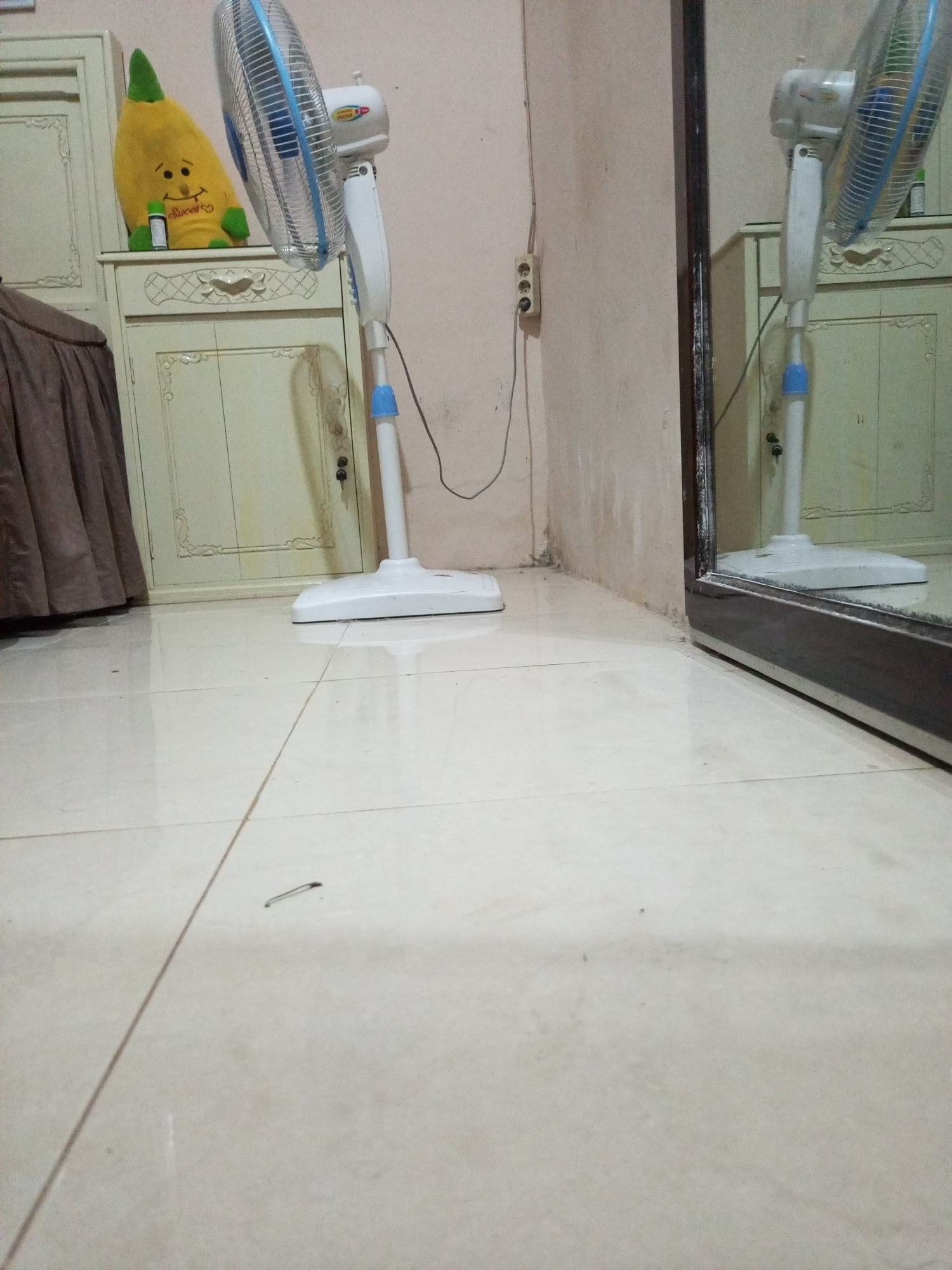 This indoor scene is located in Lhokseumawe, Indonesia, depicting a corner of a room. The floor consists of light-colored ceramic tiles with visible grout lines; a dark, elongated metallic object, possibly a hair clip, is present on the tile in the immediate foreground, with a smaller dark speck further away.
On the left, a light-colored wooden cabinet with decorative carved accents is visible. The cabinet has at least two doors, one of which has a key inserted into its lock. Resting on the cabinet's top surface are a yellow, pear-shaped plush toy with a smiling face and "Sweet" inscribed on its body, positioned next to a small, dark bottle with a white cap. To the far left, a portion of dark brown pleated fabric is visible, possibly a curtain or bed skirting.
A white standing fan, featuring a blue front grille accent and "SANNOO PROTEK" branding on its motor housing, is positioned centrally. Its power cable extends to a triple electrical outlet mounted on the light-colored wall to the left. The light-colored walls exhibit areas of grime and discoloration, particularly near the floor line. To the right, a large, dark-framed mirror stands vertically, reflecting the fan, a portion of the cabinet, and the plush toy. No individuals are present in the scene.