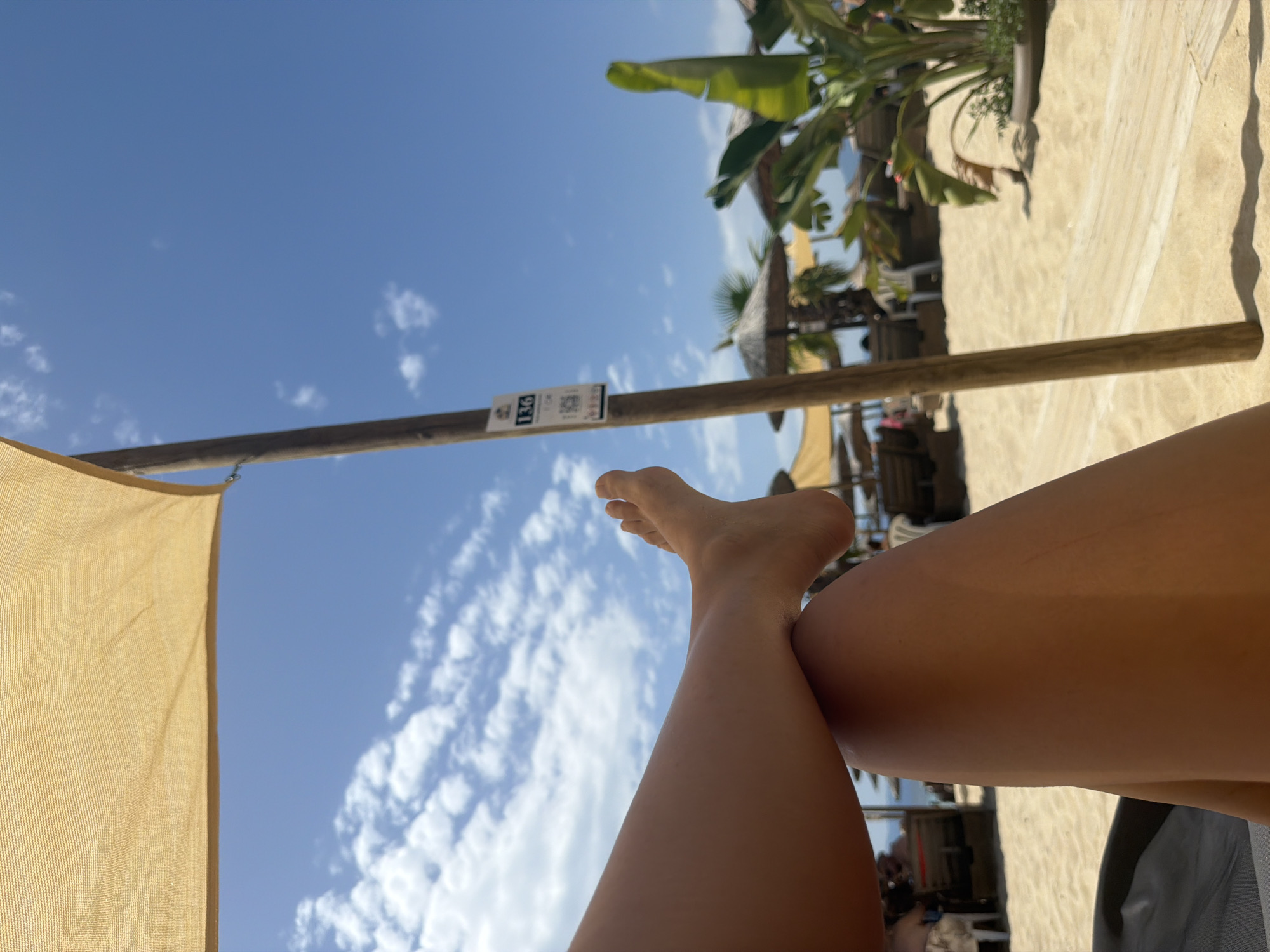 The image shows a person's legs and feet resting on a lounge chair on Ofrynio Beach, Greece. The beach is sandy, with some wooden walkways and beach umbrellas visible in the background. There are also palm trees and the sea in the distance. The sky is blue with some white clouds. A wooden pole with a sign indicating "136" is in the foreground. The person's skin appears tanned, suggesting a sunny day.