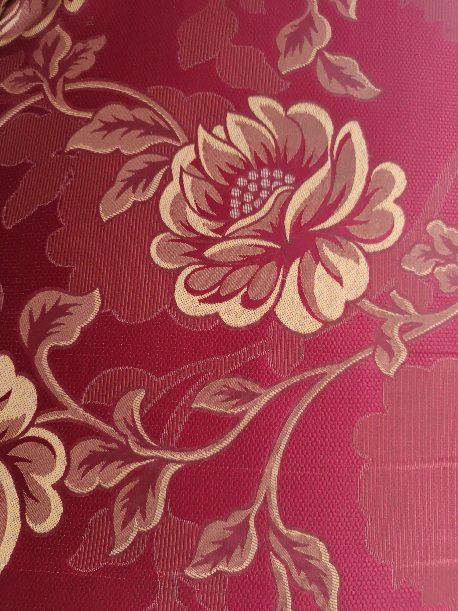 This is a close-up, full-frame, eye-level shot of a red fabric with a floral pattern. The pattern features large, stylized flowers and leaves in shades of red, gold, and beige. The fabric appears to be a brocade or jacquard weave, with raised textures creating the design. Vertical lines are visible in the red background. There are no people or other objects present in the image, nor is there any indication of a location in Gawanan, Indonesia.