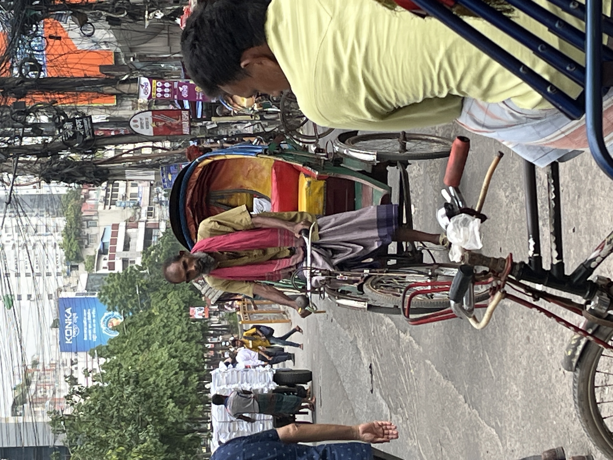 The image depicts a street scene in Dhaka, Bangladesh. In the foreground, a man is pedaling a cycle rickshaw. The rickshaw has a colorful canopy, and the man is wearing a tan shirt and a lungi. A seated man with a yellow shirt is in the foreground. In the background, a street is crowded with people and other vehicles. There are tall buildings on either side of the street. A billboard with the word "KONKA" can be seen in the background. Power lines are visible overhead.
