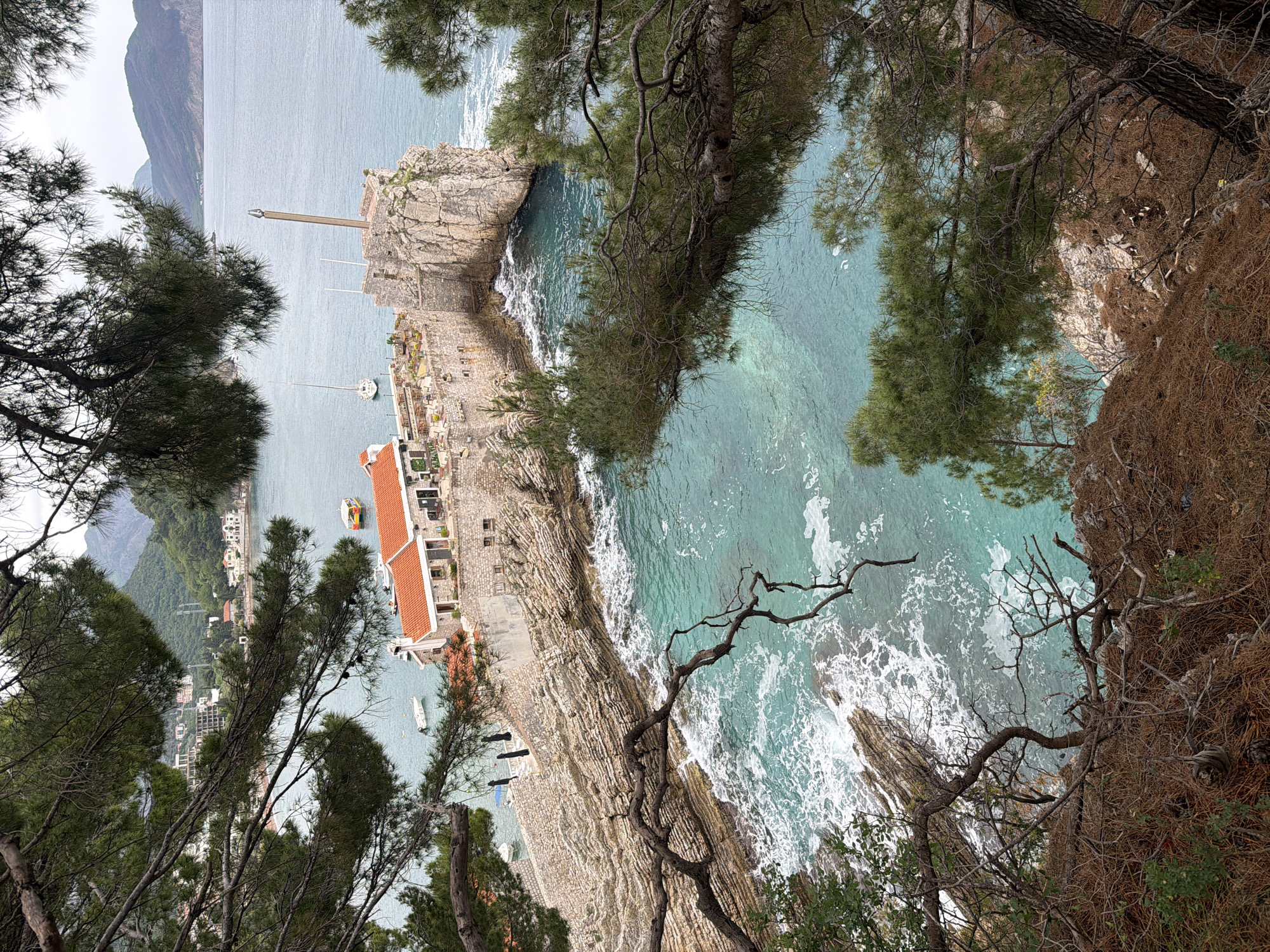 The image shows a view of the coastline in Petrovac centar, Montenegro. The camera is positioned from a height, looking down at the sea and the architectural structures along the coast. A rocky cliff descends into the turquoise waters with white foam, indicative of wave action. Trees with dense foliage frame the edges of the view, partially obscuring the scene. A building with a red tiled roof and stone walls is visible along the coast. The sea extends to the horizon, with a mountain range in the distance. A pole stands on a rocky promontory in the sea. Boats are seen in the water close to the shore.