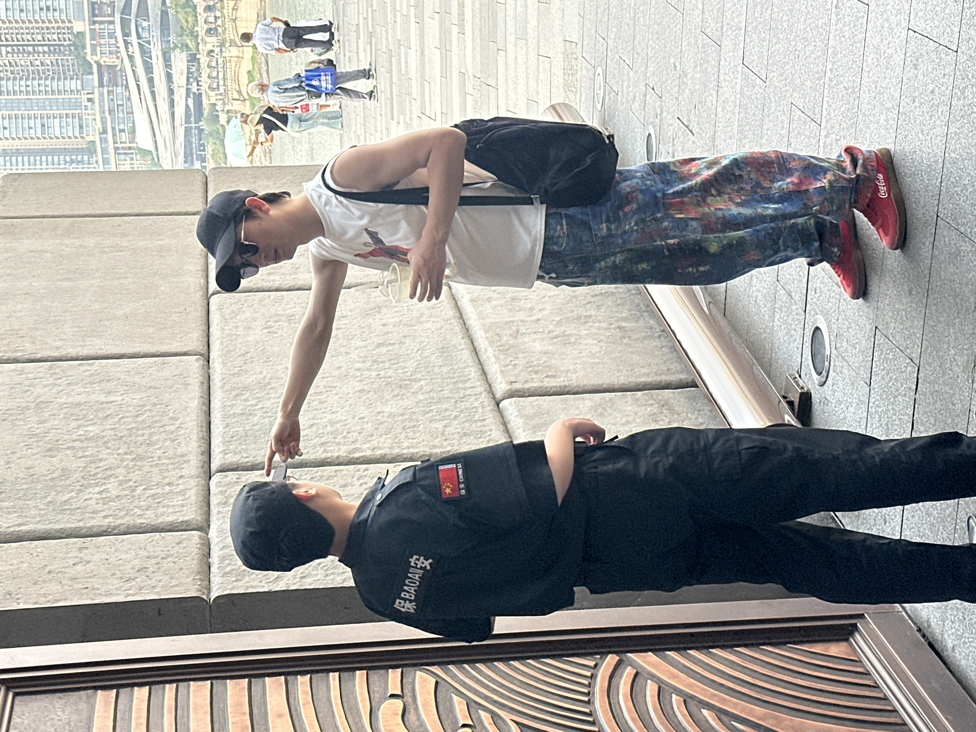 A man wearing a white tank top, black baseball cap, sunglasses, and colorful jeans points at the cap of a security guard who is lying down. The guard is wearing a black uniform with the word "BAOAN" and a Chinese flag patch on the sleeve. The scene is near a building with a decorative copper door and a stone facade. In the background, there is a view of a waterfront area with buildings and a bridge, likely within the Yuzhong District, China. Other people are walking in the background.