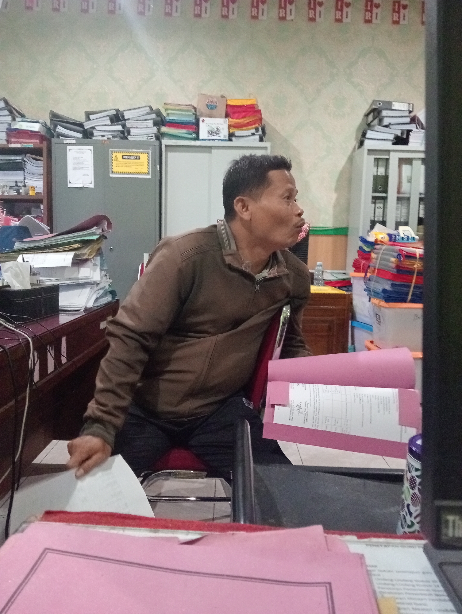 A middle-aged Indonesian man of East Asian descent is seated at a desk in what appears to be an office environment. He is wearing a brown zippered jacket over a dark shirt and blue jeans. His lips are pursed as if he is speaking or whistling. He is holding a white sheet of paper in his left hand, and his right hand is resting on the armrest of his chair.
The desk is cluttered with various documents, folders, and office supplies. There is a black tissue box, a roll of white paper towels, and a black computer monitor. Behind the man, there are several filing cabinets, shelves filled with folders, and stacks of papers. The walls are decorated with a patterned wallpaper, and there are some red and white banners hanging from the ceiling.
The overall impression is of a busy, organized workspace, likely in a government office or a business establishment in Demak, Indonesia. The lighting is somewhat dim, creating shadows on the man and the surrounding objects.