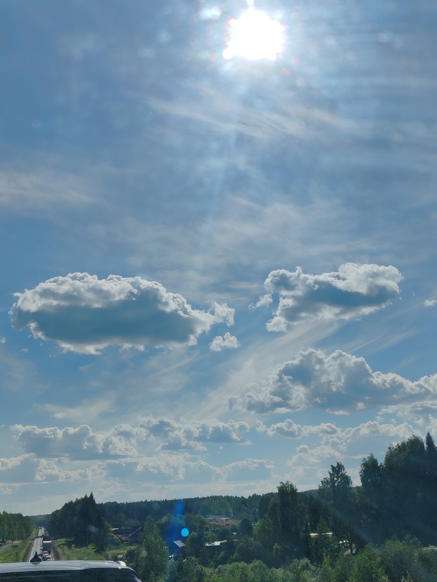 A bright, high-angle outdoor shot captures a sunlit sky with scattered clouds above a rural landscape. The sun, positioned in the upper center, is intensely bright and causes lens flare, creating a halo effect and scattering light rays across the sky. Wispy cirrus clouds are visible, interspersed with larger, cumulus clouds that cast shadows, indicating a dynamic weather pattern.
Below the sky, a dense, dark green forest stretches horizontally across the frame, forming a natural backdrop. Within the forest, small structures and buildings are partially visible, suggesting a scattered rural settlement. A road, lined with trees and a few vehicles, cuts through the scene from the left foreground towards the distant horizon. The presence of traffic implies this is a road leading to or from a populated area. The overall impression is one of a rural environment on a sunny day.
The location is specified as the village of Vylgort, Russia.