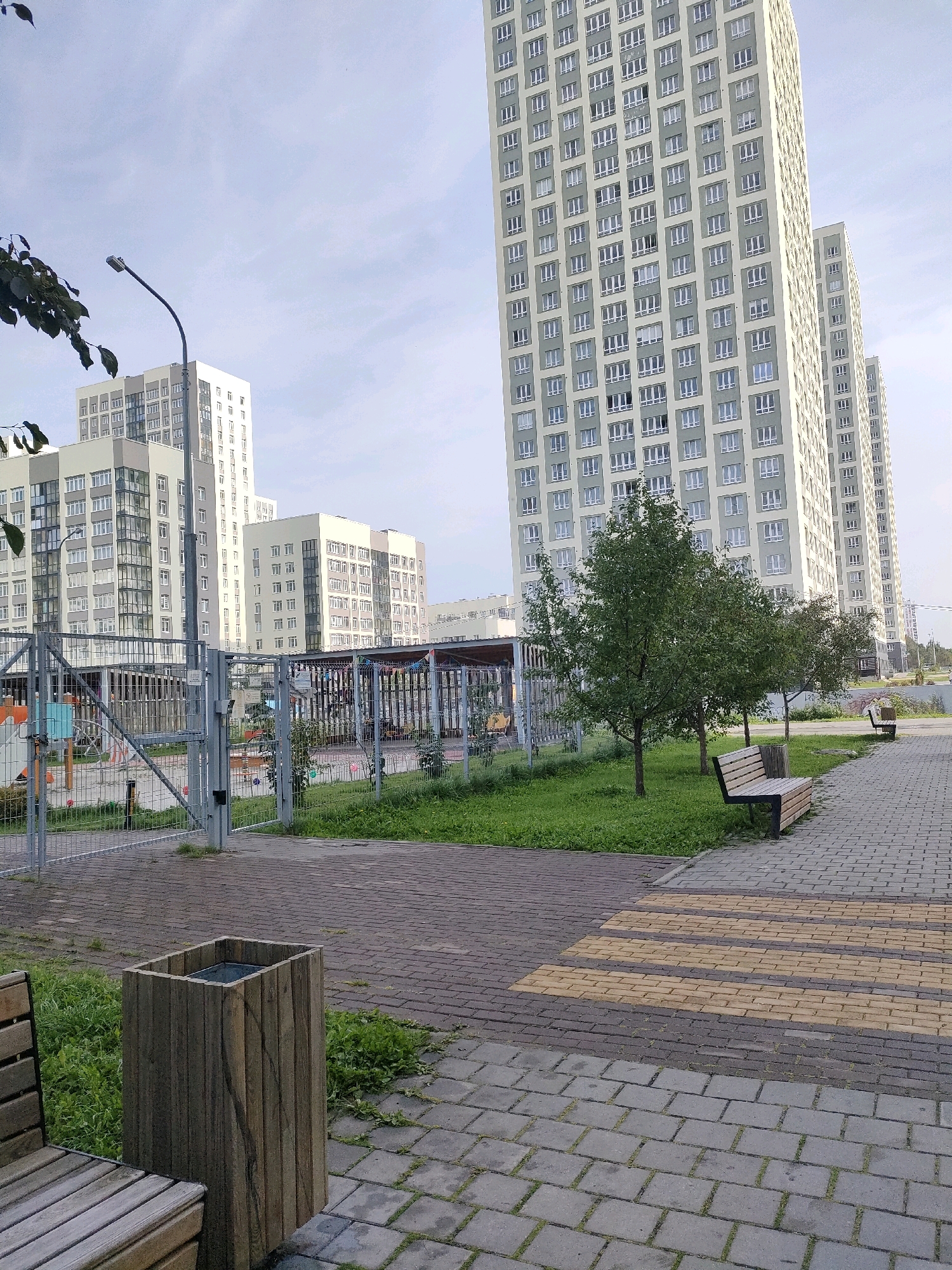 The image captures an outdoor scene in a residential area, likely in the city of Yekaterinburg, Russia, given the style of the apartment buildings. In the foreground, a paved walkway made of grey and yellow interlocking bricks leads towards the viewer. To the left of the walkway, a wooden trash receptacle stands next to a portion of a wooden bench. Beyond the walkway and trash receptacle, a grassy area is visible, bordered by a metal fence. Within the fenced area, playground equipment, including a slide and swings, can be seen. Several trees are planted in the grassy area. Behind the fence and trees, a cluster of modern, multi-story apartment buildings dominates the background. These buildings are characterized by their light-colored facades, numerous windows, and a repeating geometric pattern. A street lamp is positioned to the left of the buildings. The sky above is overcast with soft clouds. The scene appears peaceful and devoid of human activity.
