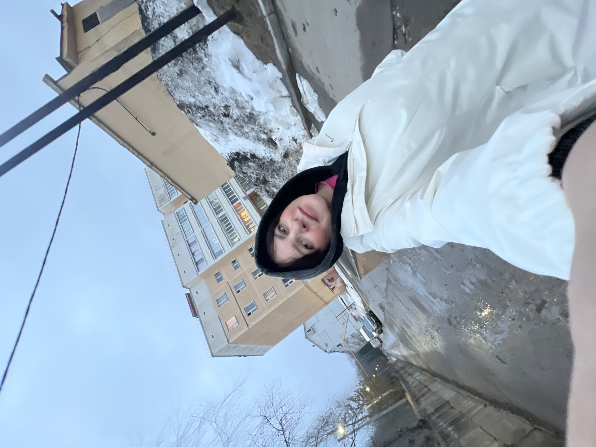 A woman wearing a white puffer jacket with a black hood is centered in the frame. Her face is visible beneath the hood. Behind her, a city street scene unfolds. A tall, multi-story building, likely an apartment complex, stands prominently in the background. Patches of snow are visible near the building, suggesting a cold climate. A wet street, reflecting the sky, indicates recent precipitation. Bare trees are visible in the lower portion of the image. Two black electrical wires stretch diagonally across the top left corner of the frame. The location is likely in the city of Noviy, Russia.