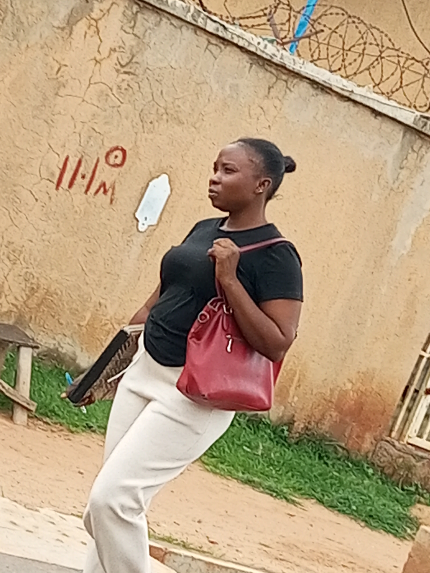 A woman is walking on a sandy path near a wall with graffiti and barbed wire. She is wearing a black t-shirt and light-colored pants. She is carrying a red bag over her shoulder and a dark-colored folder in her left hand. The environment appears to be outdoors, possibly in a residential or commercial area within the city of Gombe, Nigeria, given the style of the wall and the general appearance of the surroundings. The wall has what appears to be chalk or paint markings, including what looks like "11/1M" and a circular shape. There is also a white object attached to the wall that resembles a detached key or some other form of hardware. Green grass is visible on the left side of the image.