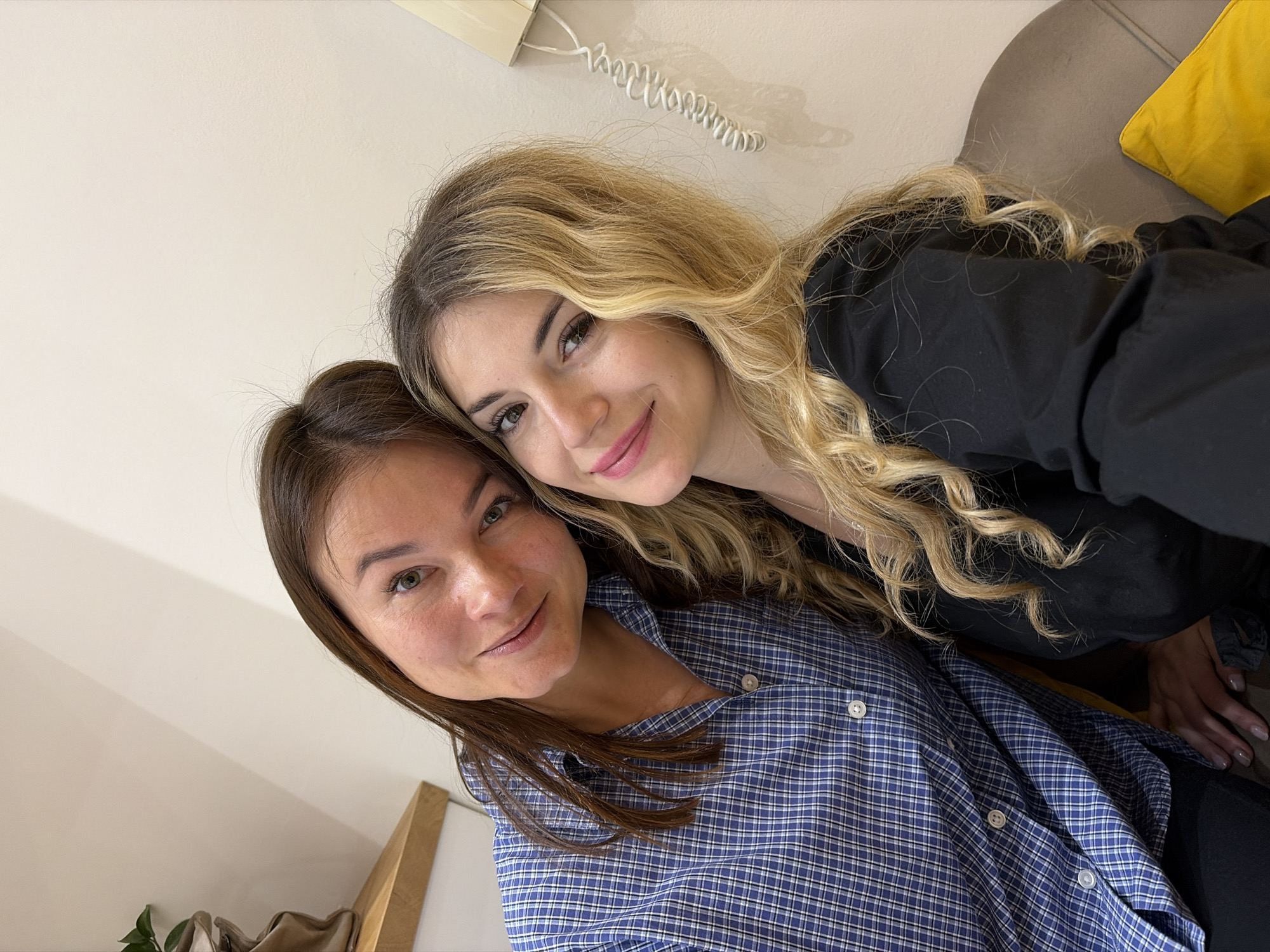 Two women are posing closely together for a selfie indoors. One woman has long blonde wavy hair and is wearing a black top. The other woman has long brunette hair and is wearing a blue plaid button-down shirt. They are both smiling. In the background, there is a white wall, a landline phone, part of a grey sofa with a yellow cushion, and part of a wooden structure. There is no information to indicate the location is in Budva, Montenegro.