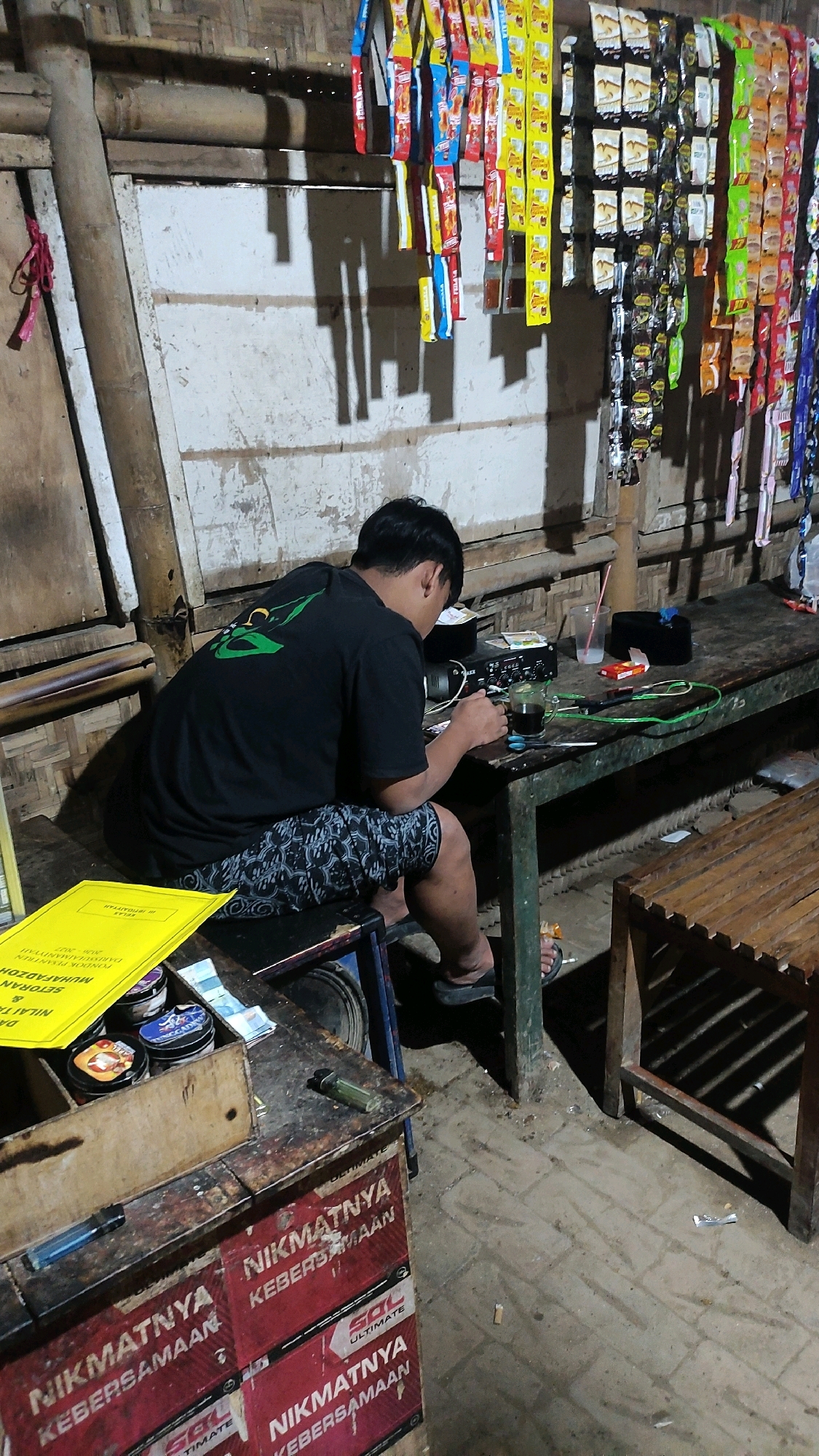 The image depicts an indoor scene, possibly a shop or a stall, in Notorejo, Indonesia. A man with dark hair is seated at a rustic wooden table, wearing a black t-shirt and patterned shorts. He is engaged with a small electronic device on the table, which also holds a glass of dark liquid, a phone, scissors, and some paper currency. The background features a wall constructed from woven bamboo or similar material, adorned with rows of hanging small packets of what appear to be snacks or condiments. The flooring is concrete, with some debris visible. To the left of the man, a wooden counter is laden with various items, including small cylindrical containers, lighters, and a yellow booklet. To the right, another wooden bench-like structure is present. The overall impression is one of a simple, functional space.