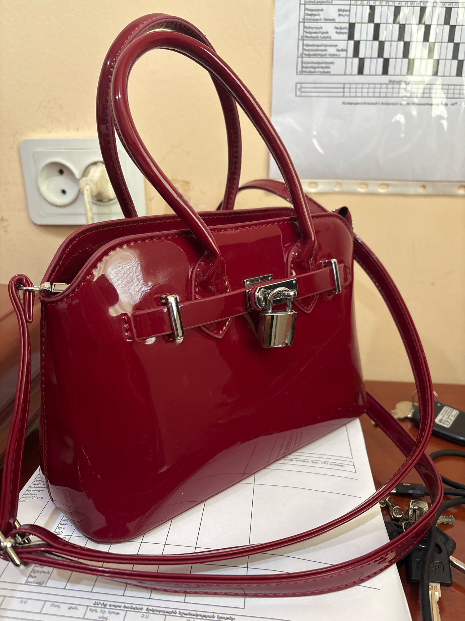 A glossy, deep red, patent leather handbag with top handles and a detachable shoulder strap sits on top of a white paper document. The handbag has a silver-toned clasp closure with a small padlock. A single electrical outlet with a white plug inserted is visible on the beige wall behind the handbag. The scene is likely indoors, given the presence of the electrical outlet and the paper document. The image does not provide any specific visual cues to indicate a location within the city of Ijevan, Armenia.