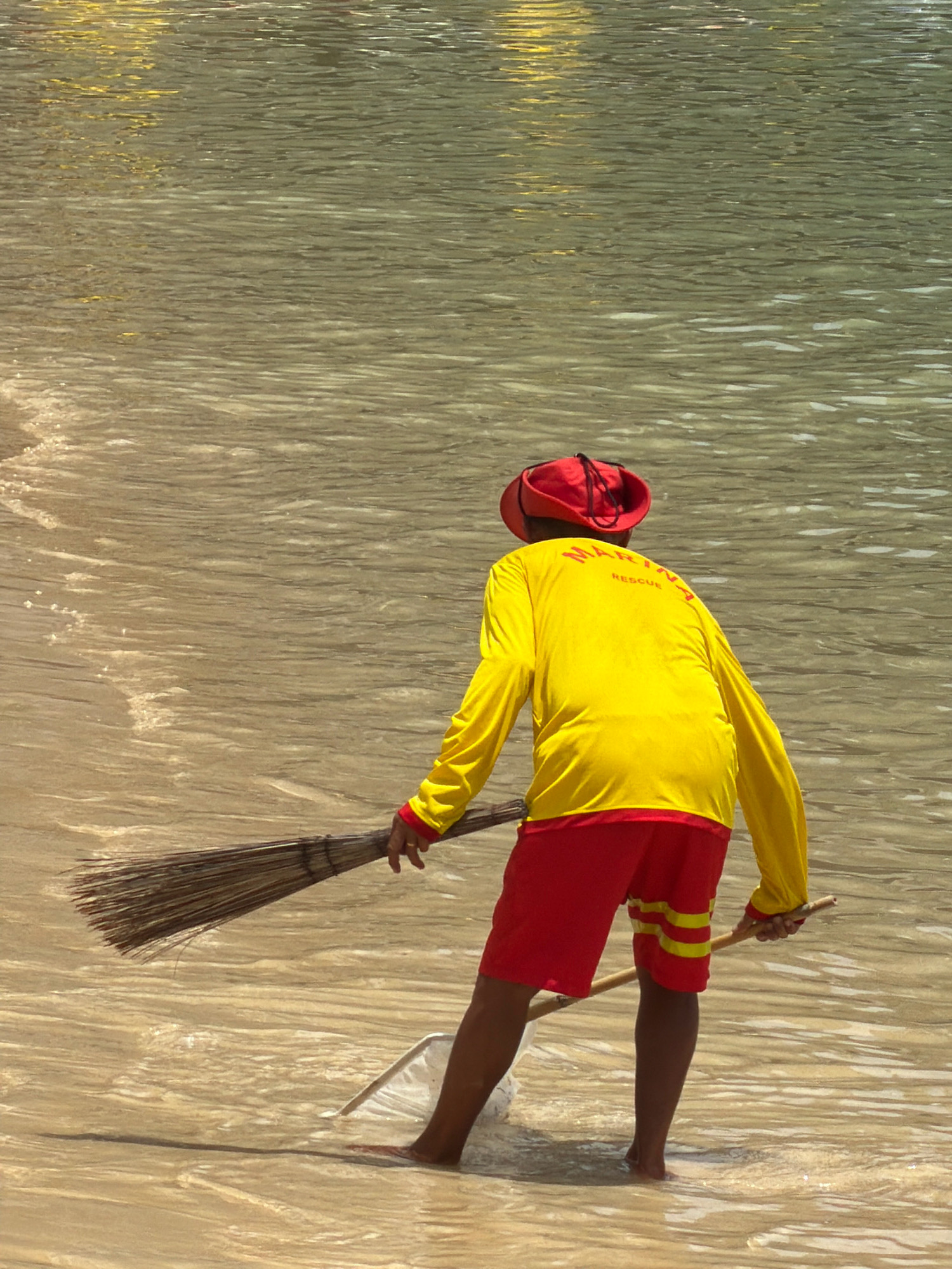In Nha Trang, Vietnam, on a sunny day, a lone individual, dressed in a bright yellow long-sleeved shirt with red trim and red shorts with yellow stripes, stands in shallow, clear water near the shore. The person wears a red hat and is holding a broom with a wooden handle, sweeping what appears to be debris or seaweed in the water. A white net is partially submerged near their feet. The water is calm with ripples reflecting the sunlight, and the sandy seabed is visible. The background consists of more water with yellow reflections, suggesting a beach or coastal area. The word "RESCUE" is visible on the back of the yellow shirt.