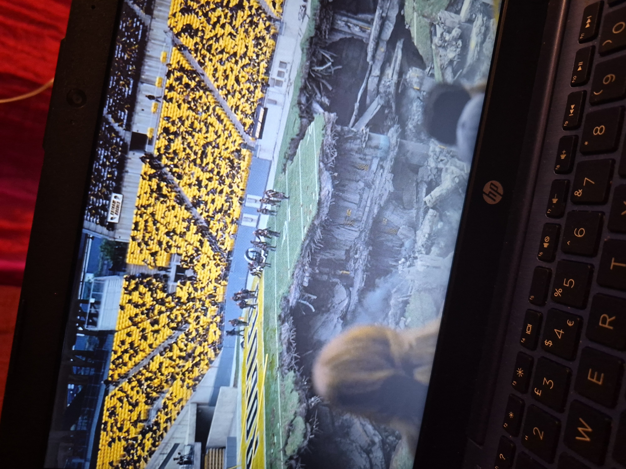 The image shows a laptop screen displaying a scene of a football stadium with a large crater in the middle of the field. The stadium seats are filled with spectators wearing yellow and black. On the field, several people appear to be walking towards the crater. The laptop keyboard is visible in the foreground. There is no information in the image to indicate the location in Sandwell, United Kingdom.