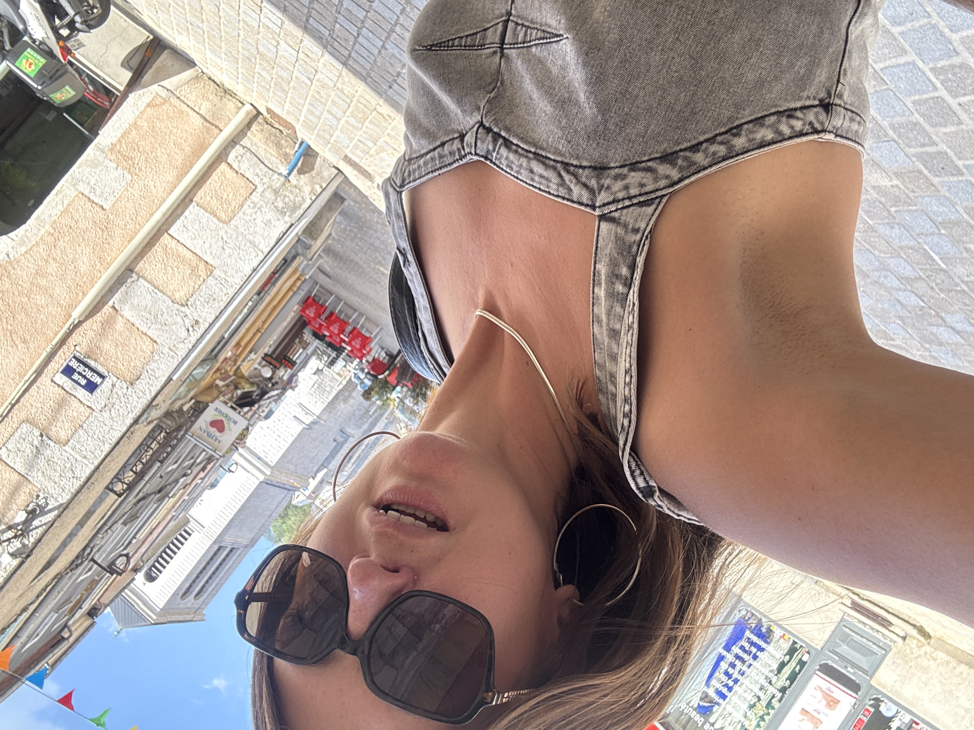 A woman, wearing sunglasses and a denim overalls, is looking up at the camera. She has blonde hair and is wearing large hoop earrings and a thin silver necklace. The background is a street scene in Saint-Claude, France, with buildings, red chairs outside a cafe, and a church tower visible. Colorful triangular flags are strung across the street.
