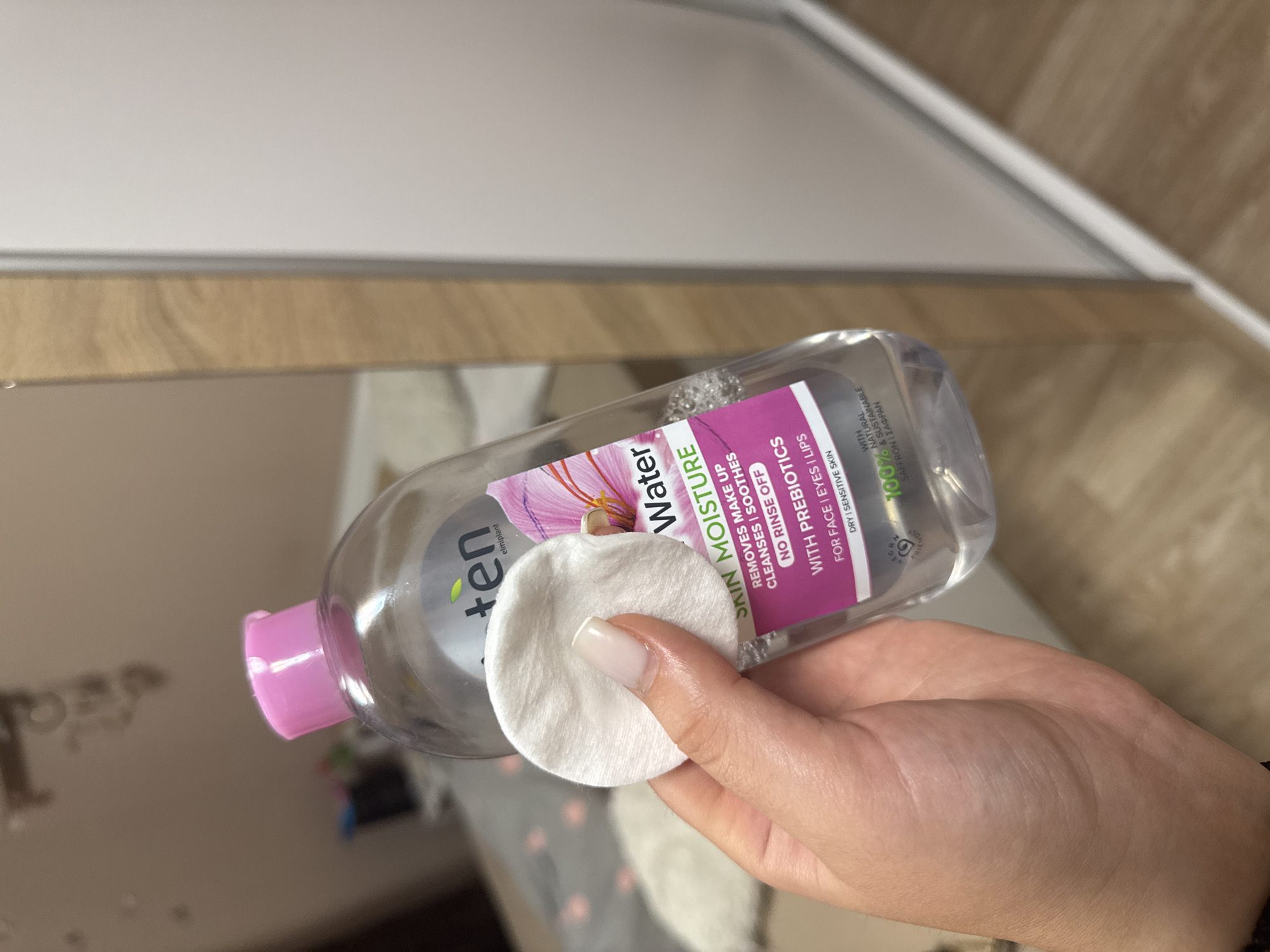 The image shows a close-up of a person's hand holding a white cotton pad in front of a clear plastic bottle of "skin moisture" micellar water. The bottle has a pink cap and a label with text in purple and green. The background is blurred but appears to be an indoor setting, possibly a bedroom or bathroom, with what looks like a wardrobe or cabinet on the right side. The overall lighting is bright. There is no indication of the location being in Rogosh, Bulgaria, or any specific city.