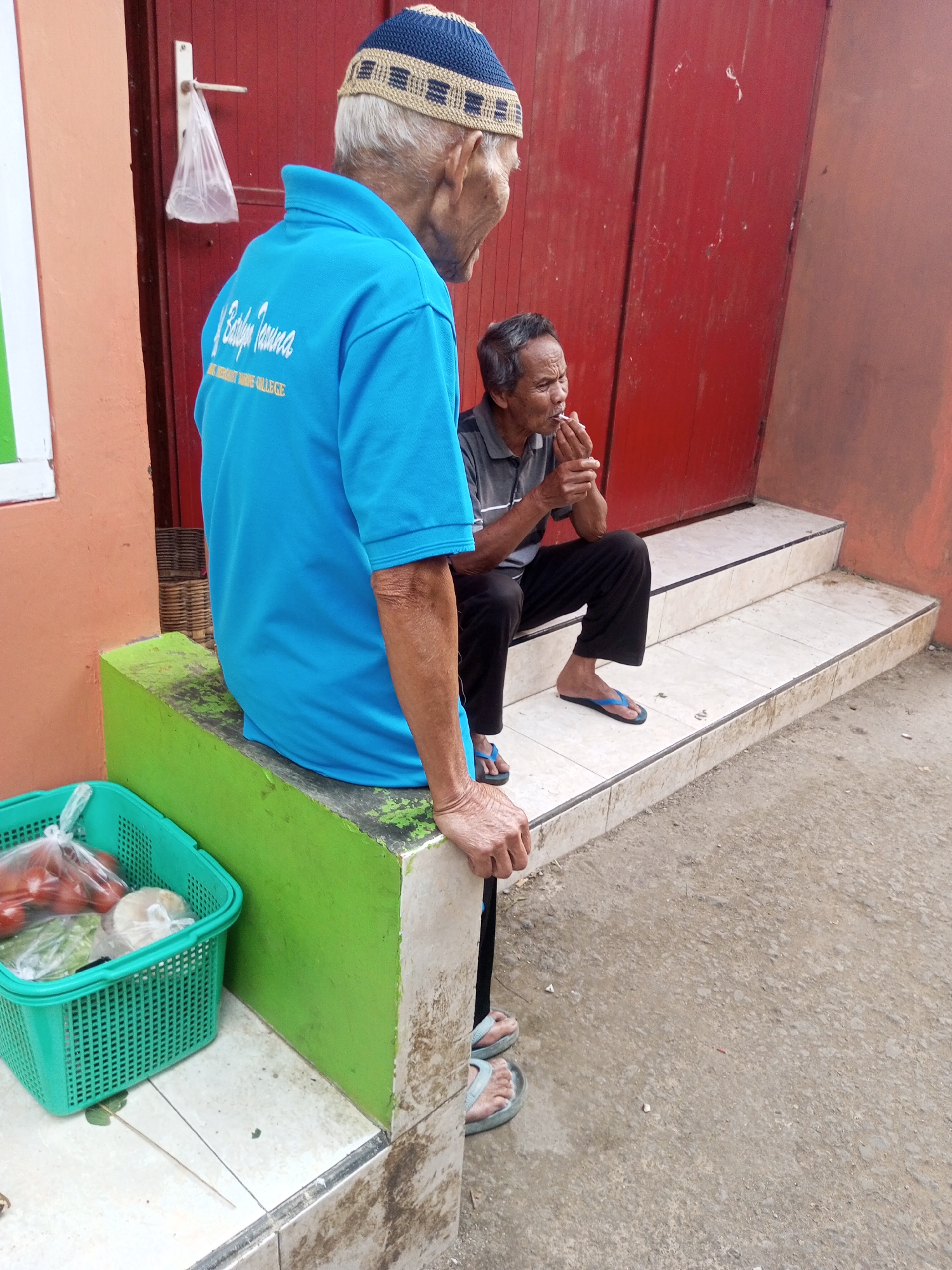 Two elderly men are sitting outside a building with a red door. The man on the left is wearing a blue polo shirt and a knitted blue and tan cap, and is looking to the right. He has his left hand resting on a green block in front of him. The man on the right is wearing a gray striped shirt and black pants, and is sitting on a tiled step with his legs crossed. He is holding a cigarette to his mouth and appears to be smoking. In the foreground, to the left of the man in the blue shirt, is a green plastic basket containing produce, including tomatoes and some leafy greens. The ground is a mixture of concrete and dirt. The building has an orange-colored wall on the left. The scene appears to be in Kalibeber, Indonesia.