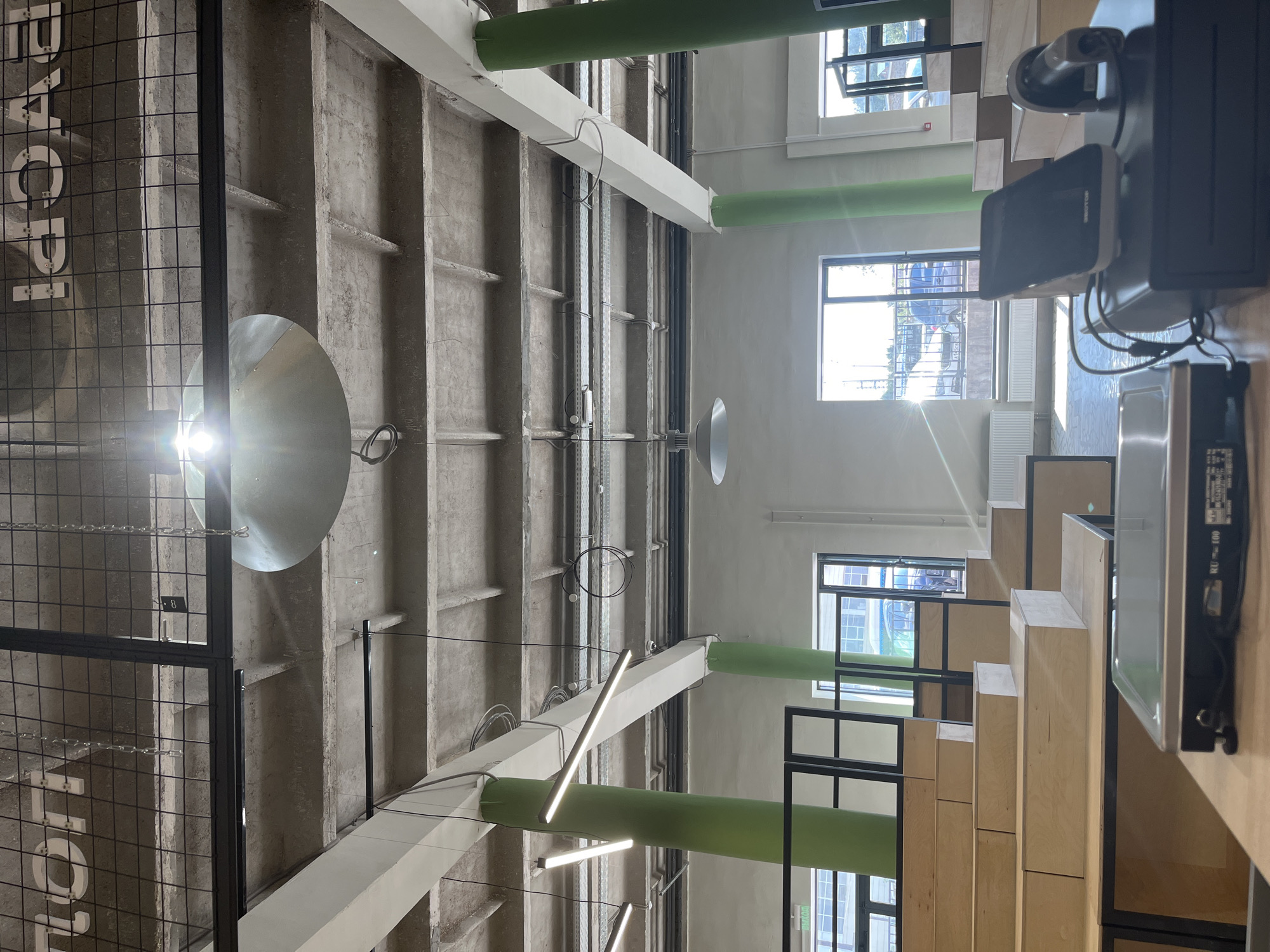 The image shows the interior of a modern marketplace or retail space, possibly a food hall, located in Ufa, Russia. The architecture features an industrial aesthetic with exposed concrete ceilings and beams, complemented by bright green cylindrical support columns. The space is well-lit, with a combination of natural light streaming through large windows and artificial lighting from linear fixtures and a large pendant lamp.
In the foreground, a retail counter is visible with a digital scale and a point-of-sale system, including a touchscreen monitor and a barcode scanner. Behind the counter, tiered wooden seating, reminiscent of an amphitheater or stadium, fills the space. The seating is constructed from light-colored wood with black metal framing.
Through the large windows, an outdoor scene is visible, showing a street or parking area with cars and some distant structures, indicating an urban environment. The overall impression is of a clean, contemporary, and functional commercial space.