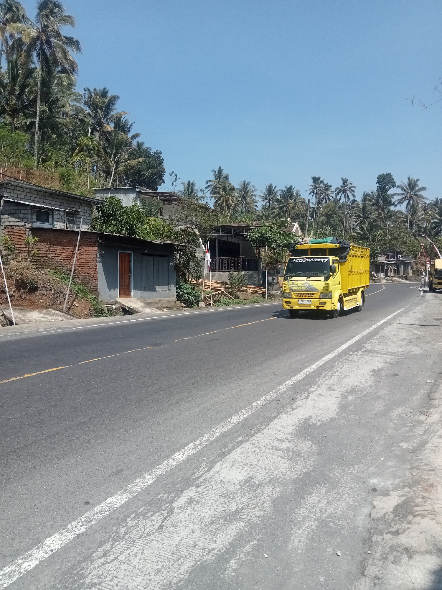 A bright yellow truck with a covered cargo bed drives on a paved road under a clear blue sky. The truck has a white stripe on its side with the text "AnginWangi" in black lettering. The truck is heading away from the camera and is on the right side of the road. To the left of the road are some low-rise buildings, some made of brick and others with grey siding. A dirt embankment with vegetation rises behind the buildings. Coconut palm trees are visible in the background, lining the top of the embankment and further into the distance. The road has a yellow line marking the center and a white line on the right edge. Some white markings on the road surface suggest previous wear or repairs. The overall scene appears to be in a rural or semi-rural area with tropical vegetation.