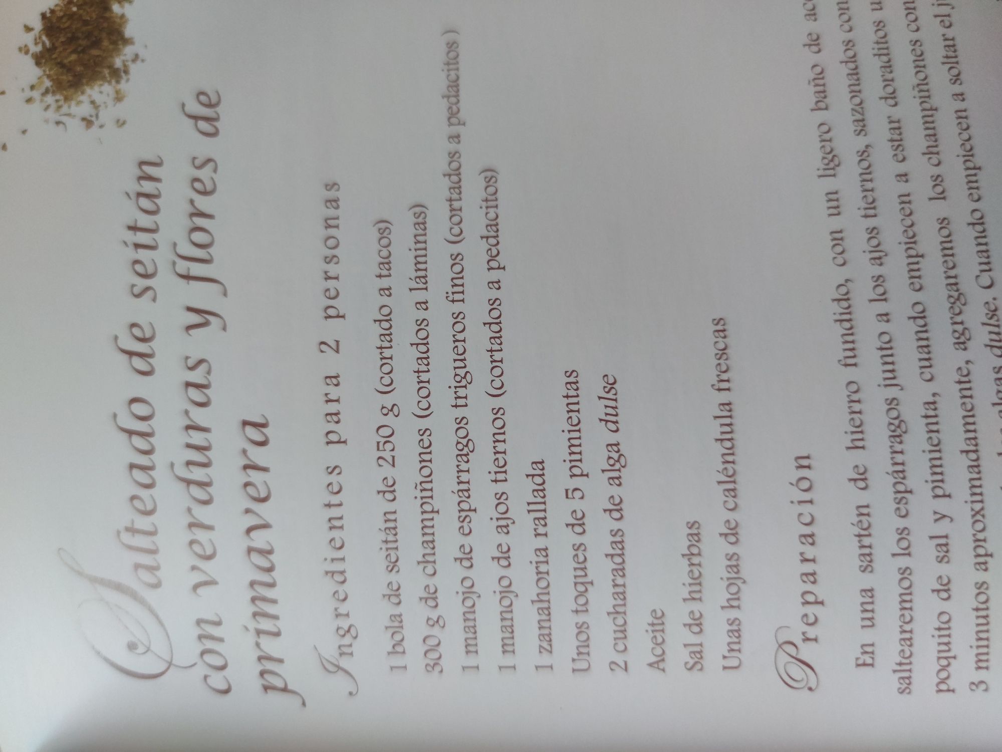The image is a close-up photograph of a page from a recipe book. The text is written in Spanish in a brown font. The title of the recipe is "Salteado de seitán con verduras y flores de primavera" (Seitan stir-fry with spring vegetables and flowers). Below the title, a list of ingredients is provided for 2 people, including seitan, mushrooms, thin asparagus, young garlic, grated carrots, 5 peppercorns, dulse seaweed, oil, herbal salt, and calendula leaves. The recipe then continues with the "Preparación" section which gives instructions for cooking. In the upper left corner of the photo are some dried herbs.