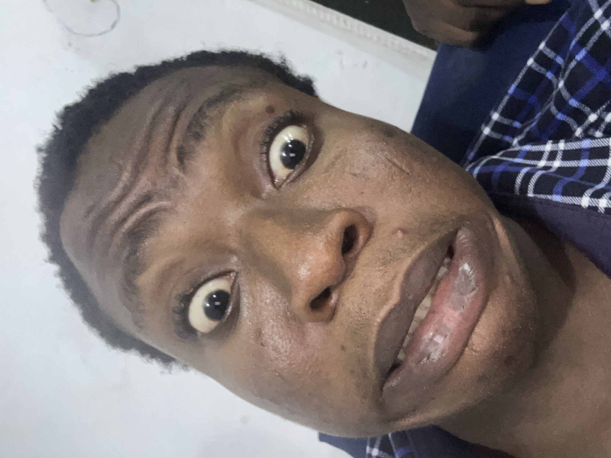 A close-up, tilted, color photograph shows a dark-skinned person from the chest up. The person has short, dark hair. The eyes are wide and focused. A plaid shirt with blue and white squares is visible around the neck and chest. The background is a plain white wall.