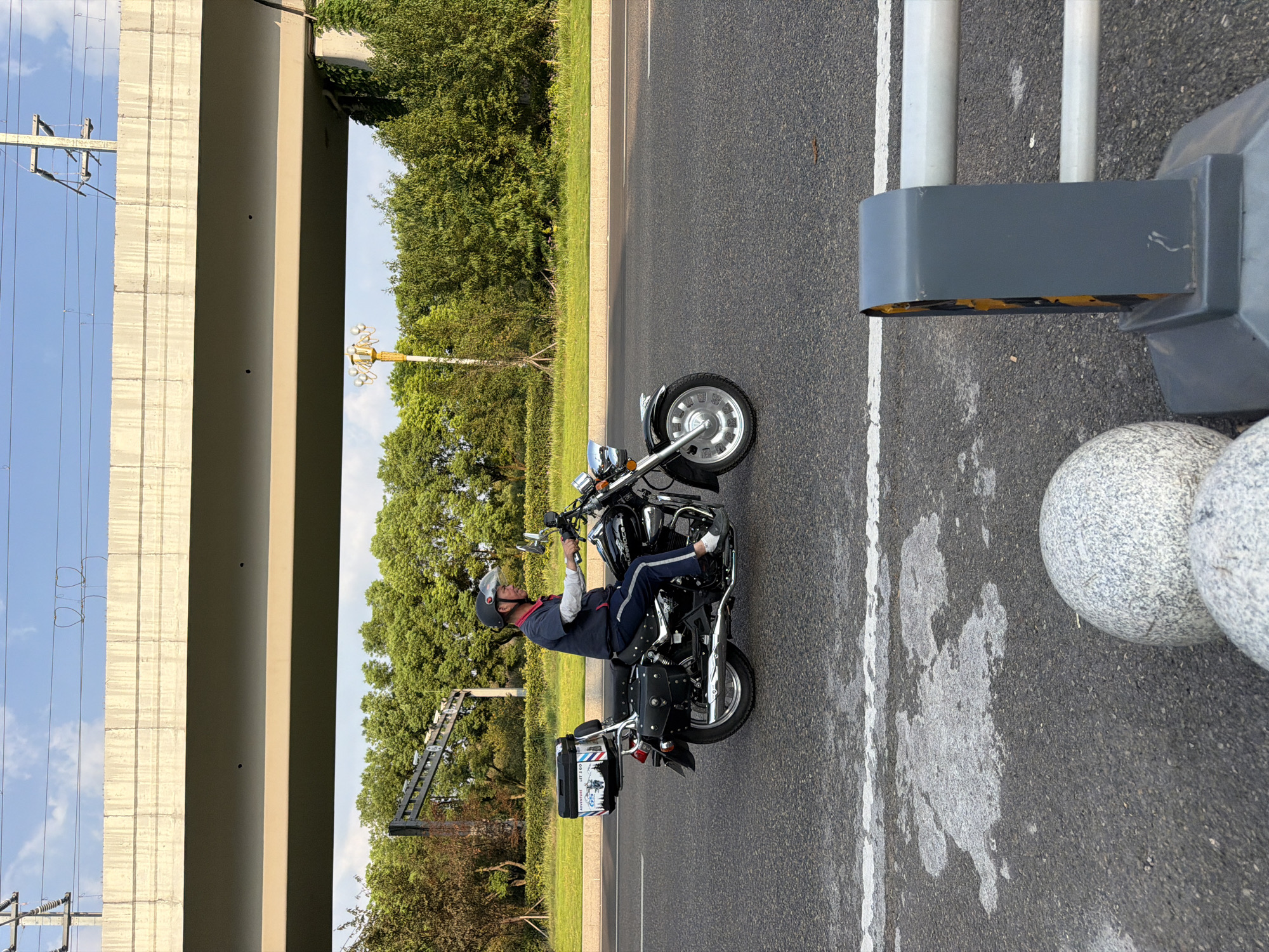 An outdoor, eye-level, medium shot shows a man riding a black cruiser motorcycle on a paved road under a concrete overpass in Jiangjin, China. The man, wearing a blue shirt, blue pants, a black helmet, and white shoes, is riding to the right of the frame. He is looking forward and has his hands on the handlebars. The motorcycle has a black storage box on the back. The road has white lane markings. In the background, there are trees, a yellow streetlamp, and overhead power lines. In the foreground, there are two large spherical bollards and a grey traffic barrier. The sky is blue with some clouds. The lighting suggests it is daytime, likely late afternoon due to the long shadows.