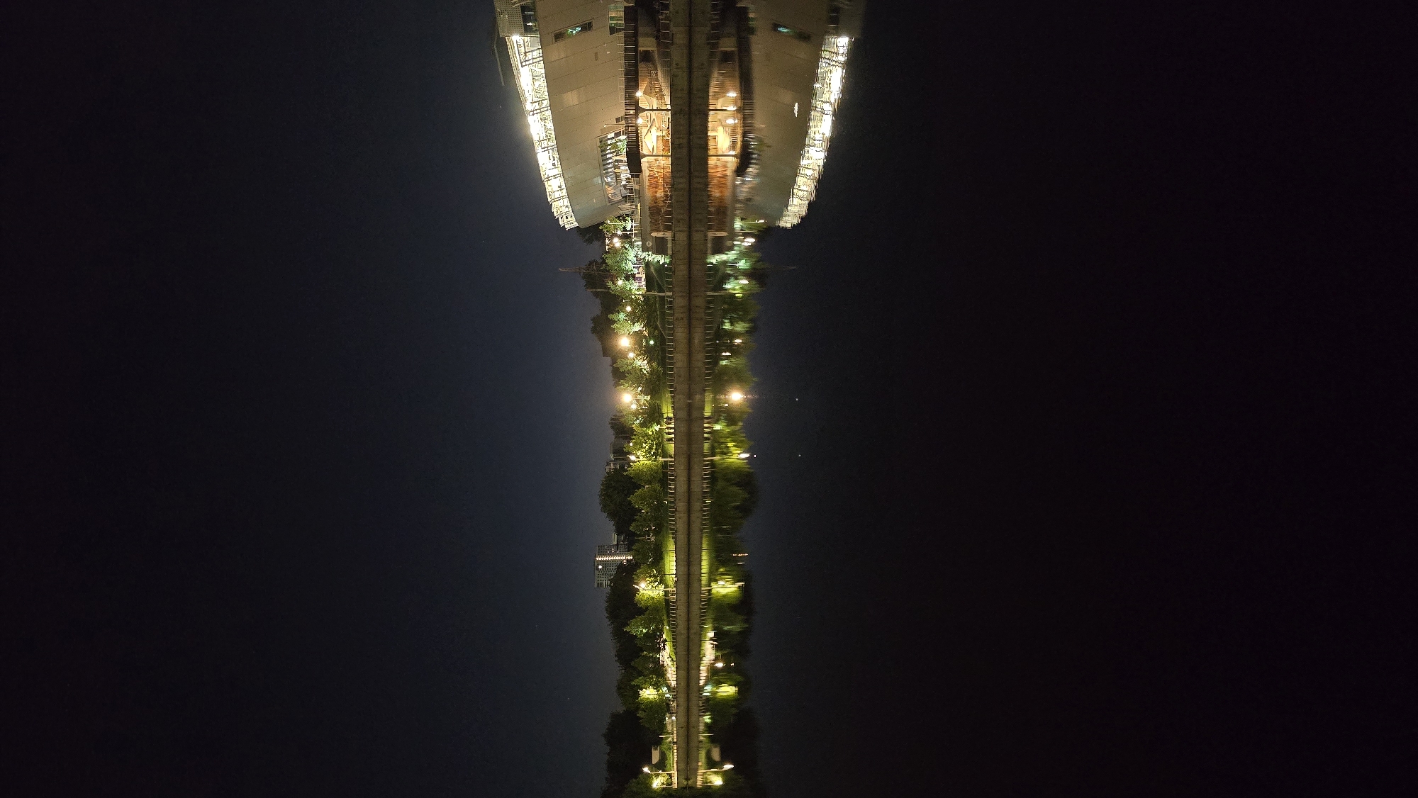 The image shows a nighttime view of the embankment of the Dnipro River in Kyiv, Ukraine. A modern, illuminated building with a glass facade, likely a cultural or entertainment venue, is prominent on the right side of the scene. Along the riverbank, trees are lit by streetlights, creating a reflective pattern on the dark water. A few people are visible walking along the embankment. The overall atmosphere is serene and urban, illuminated by artificial lights against the dark night sky.