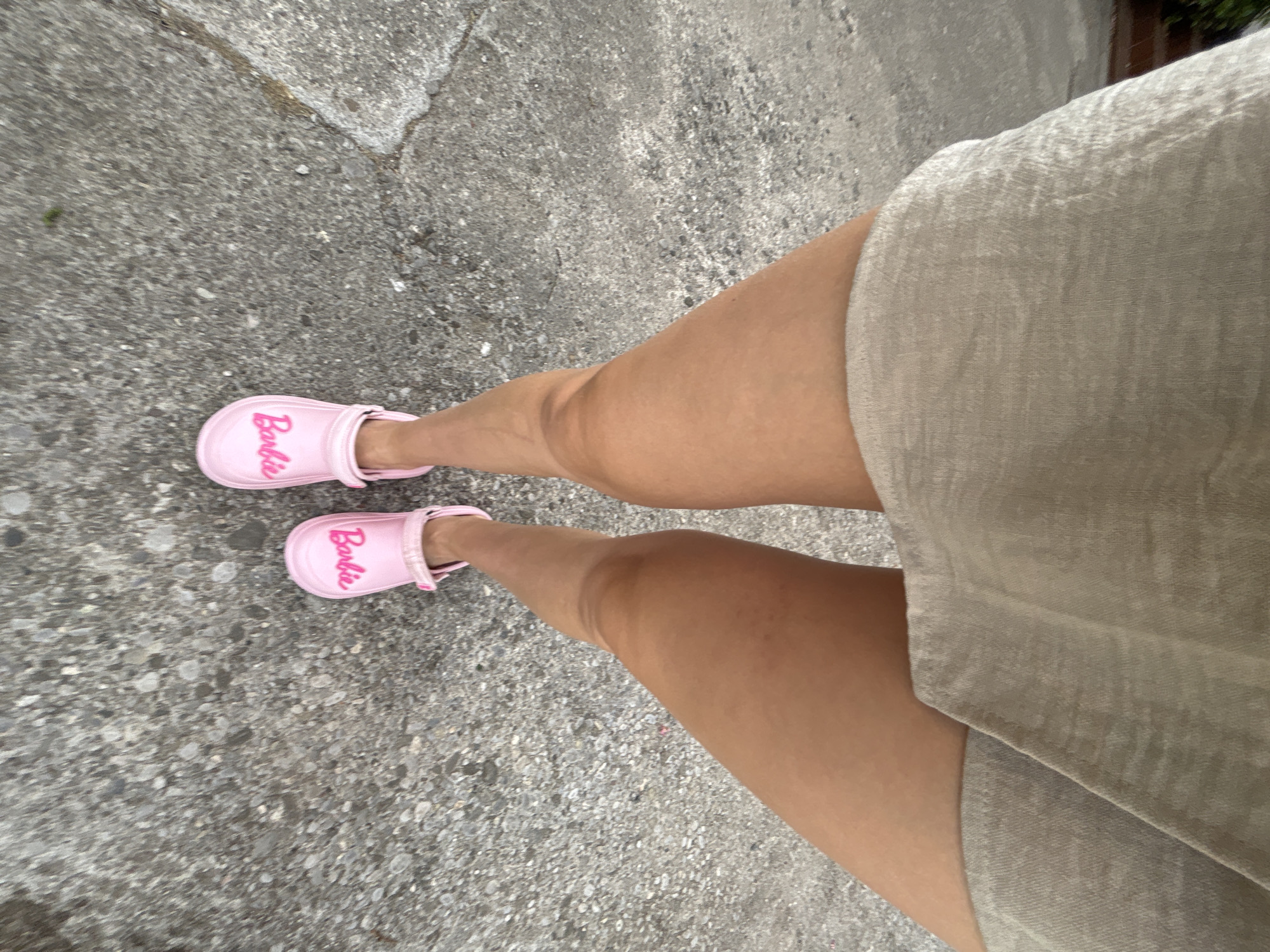 A low-angle, full-shot, eye-level, medium-wide photograph shows a person's legs from mid-thigh down, standing on a paved surface that appears to be a sidewalk or street. The person is wearing a light-colored, knee-length A-line dress or tunic and bright pink "Barbie" branded clogs. The clogs are the most visually striking element of the footwear, featuring the word "Barbie" in a stylized pink font on the front. The person's legs are tanned. The paved surface is grey, textured, and shows signs of wear and tear, with visible cracks and small stones embedded in it. The background is blurred, suggesting a shallow depth of field, but appears to be an outdoor urban setting with some greenery visible on the left side. The lighting suggests it is daytime, possibly overcast. The location in Budva, Montenegro is not discernible from the visual information provided in the image.