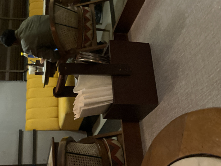 The image captures an indoor scene, likely a restaurant or cafe, with a warm, somewhat dim ambiance. The perspective is rotated 90 degrees clockwise, showing the scene from an elevated angle.
In the mid-ground, a person, identifiable as a man with dark hair, is seated at a dark wooden table. He is wearing a dark green long-sleeved shirt and appears to be looking downwards, possibly at something on the table or his phone. He is seated on a wooden chair featuring a cane back and a cushion with a geometric pattern in red and white tones.
To the left of the man and extending along a wall is a prominent yellow padded booth or bench, providing a vibrant color contrast to the otherwise muted tones. On the table, various items are visible, including a small bottle (possibly a condiment) and other table settings. Adjacent to the table, in the right foreground, is a dark brown wooden napkin holder, densely packed with white paper napkins. Above the napkin holder, a stack of metallic plates is resting. Another similar wooden chair with a cane back is partially visible in the bottom right corner of the frame. The floor appears to be a light, textured surface.
The lighting is soft and subdued, typical of an evening setting indoors. There are no distinct location cues within the image itself, though the provided context indicates the location is Dibrugarh, India. No visible text is discernible anywhere in the frame.