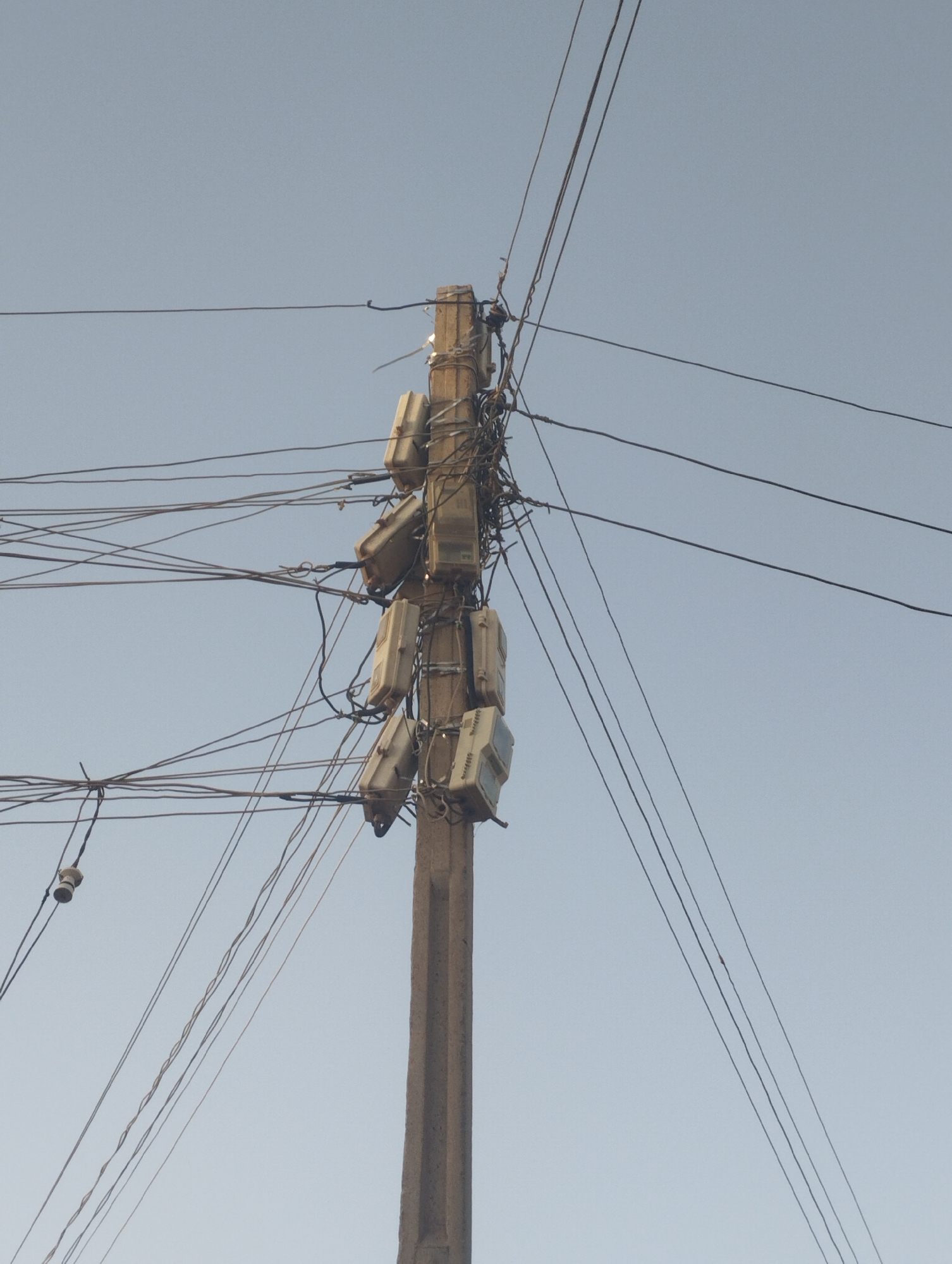 The image shows a concrete utility pole in Tumfure, Nigeria. Multiple electrical meters are attached to the upper portion of the pole, along with a dense and tangled network of electrical wires extending in various directions. The sky is visible in the background, appearing light blue. There are no people visible in the frame.