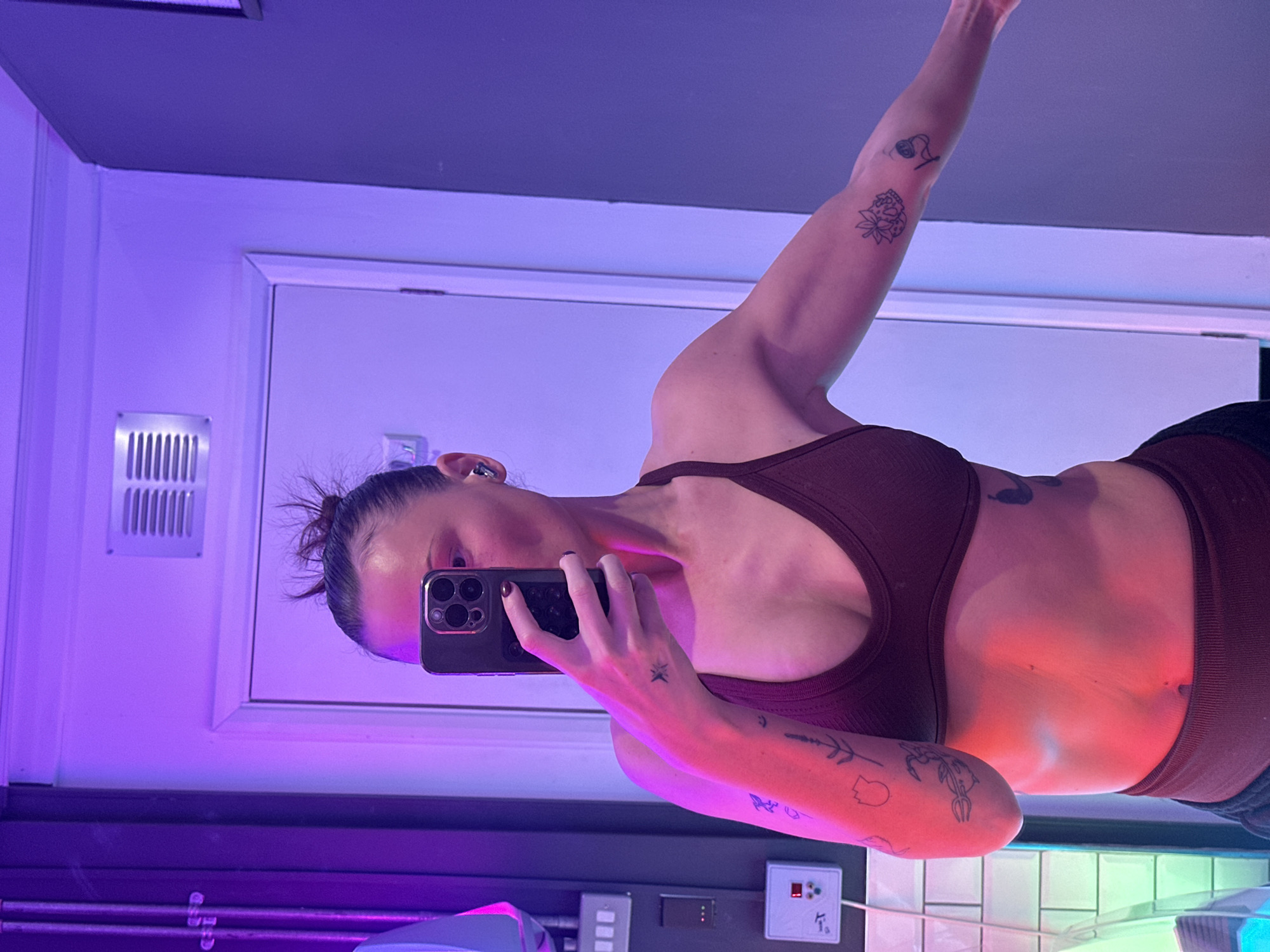 A person is taking a selfie in a room with blue and purple lighting. They are wearing a brown sports bra. Tattoos are visible on their arms and abdomen. The room has white walls, a gray ceiling, and what appears to be a tiled area near the bottom of the frame. Electrical outlets and equipment are visible in the lower portion of the photo. The specific location within Tonbridge and Malling, United Kingdom is not identifiable from this image.