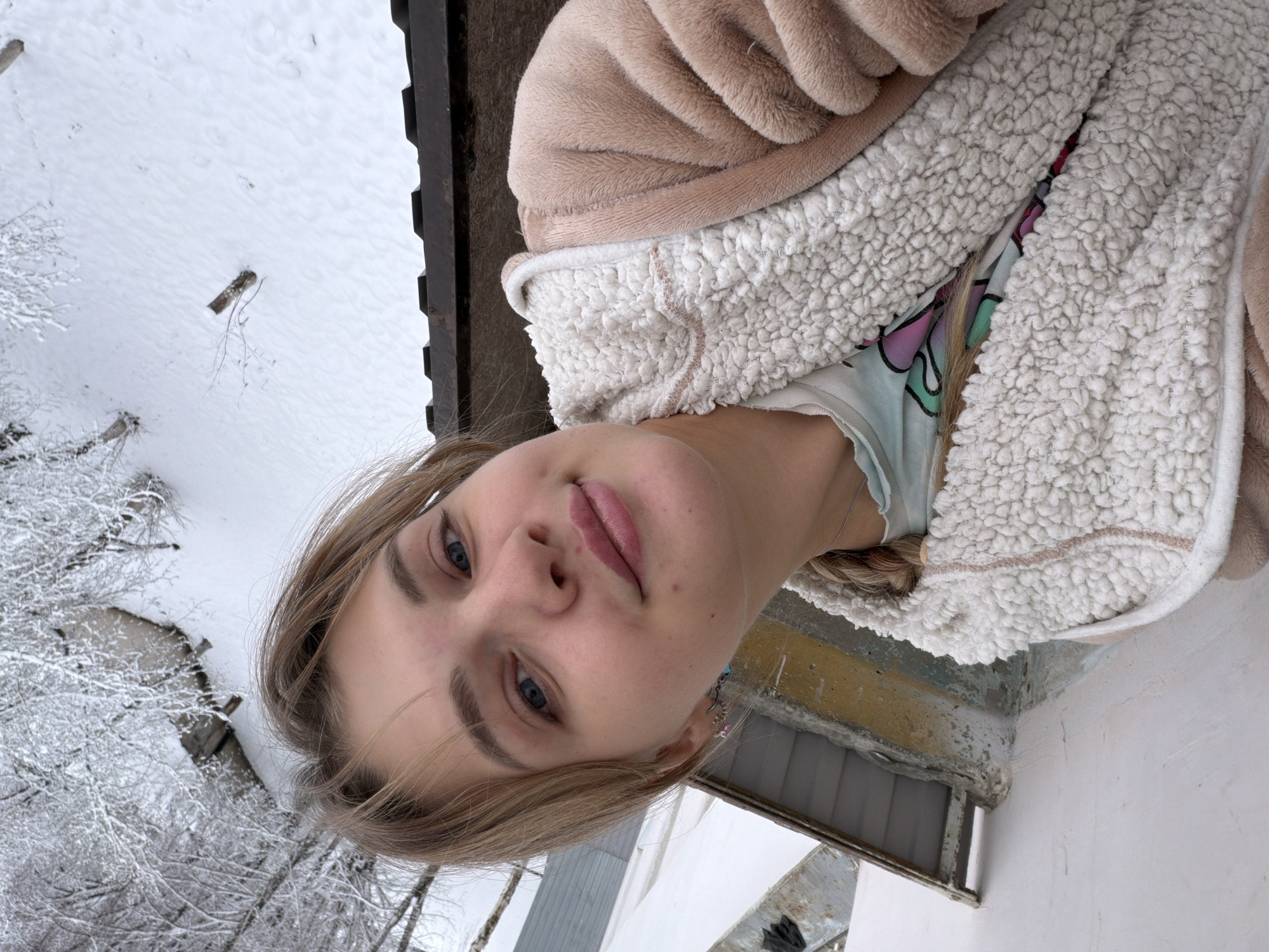 A young woman with blonde hair and blue eyes is centered in the foreground, wearing a light brown and white sherpa-lined hooded jacket over a light blue patterned shirt. She appears to be taking a selfie outdoors, possibly on a balcony or near a building. In the background, there is a snow-covered landscape, including trees and what may be a frozen body of water. The presence of snow and what appear to be snow-covered trees suggests it is winter. In the upper left, there's a structure resembling a roof or balcony. In the right and bottom of the image, a building exterior is visible. There are no specific landmarks immediately visible that would definitively indicate Naberezhnye Chelny, Russia.