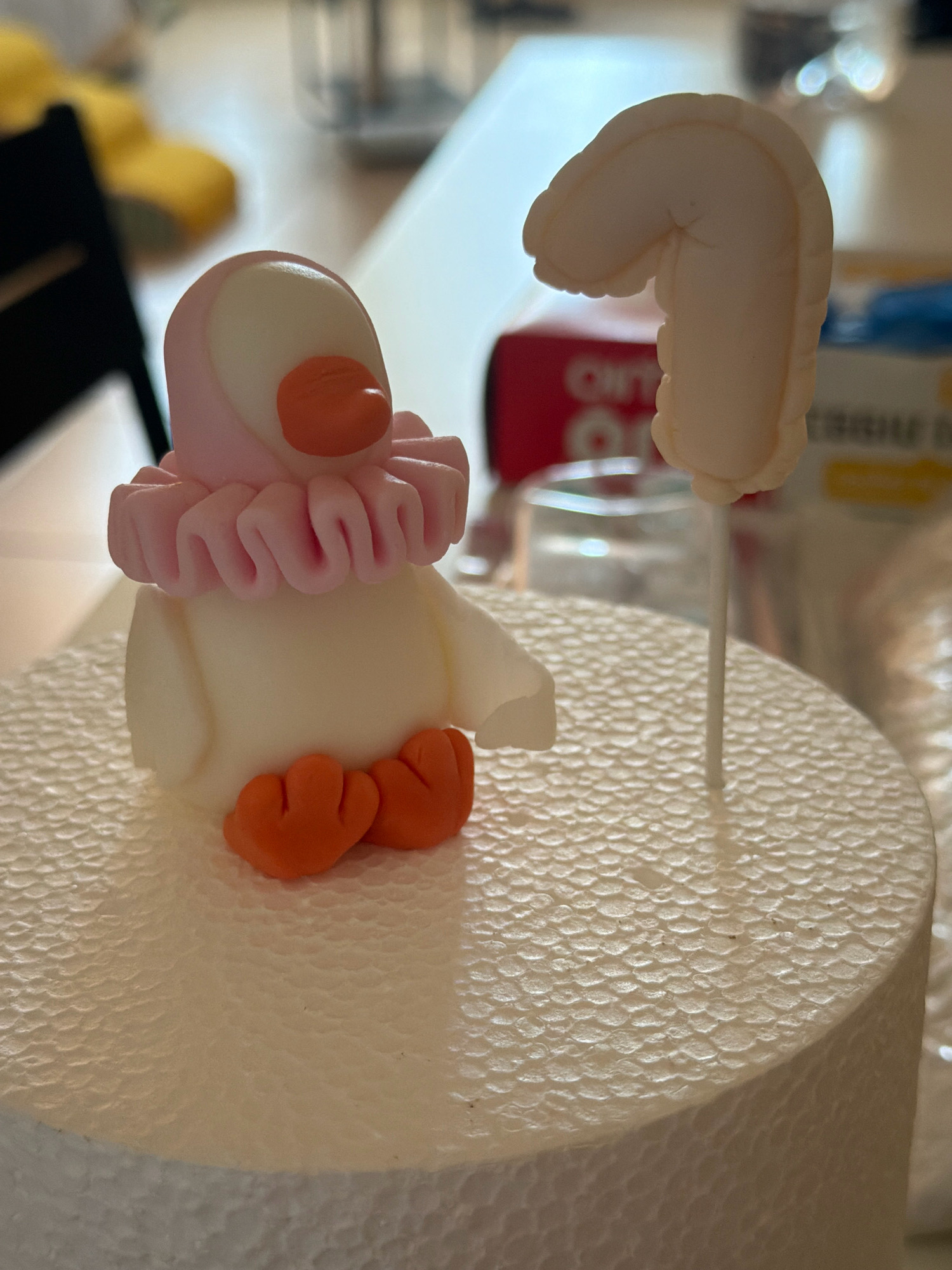 A fondant duckling with orange feet and beak, and a pink frilled collar, sits on a white textured cake base. Next to it, on a white stick, is a cream-colored fondant number "1". The background is blurred, showing what appears to be a domestic setting with kitchen items and packaging. This scene is not located in Tel Aviv, Israel, as there are no identifiable landmarks or context to suggest this location.