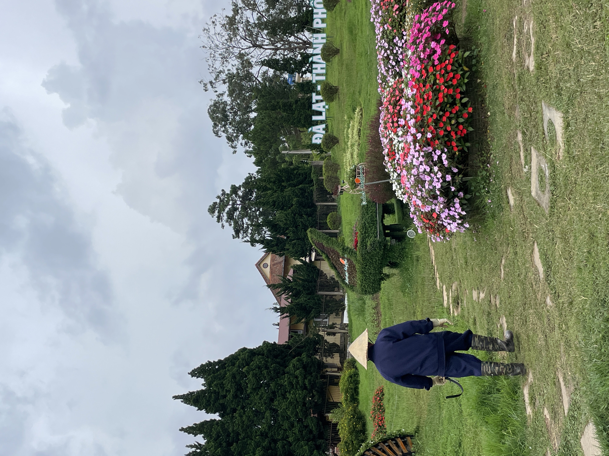 The image depicts a garden scene in Da Lat, Vietnam, characterized by its lush greenery, vibrant flower beds, and decorative topiary. In the foreground, a person wearing a conical hat, a dark blue jacket, dark pants, and camouflage boots walks away from the camera on a stone pathway. The person is holding a tool. To the right, a large bed of colorful flowers in shades of pink, red, and white is prominently displayed. In the mid-ground, a meticulously shaped topiary resembling a peacock or bird is visible, surrounded by green bushes. Further back, a building with a yellow facade and a red roof is partially obscured by tall cypress trees. Behind the building and the trees, a large sign reads "DA LAT - THANH PHO" in white lettering against a backdrop of trees and a cloudy sky. The overall impression is of a well-maintained public garden or park within the city of Da Lat.