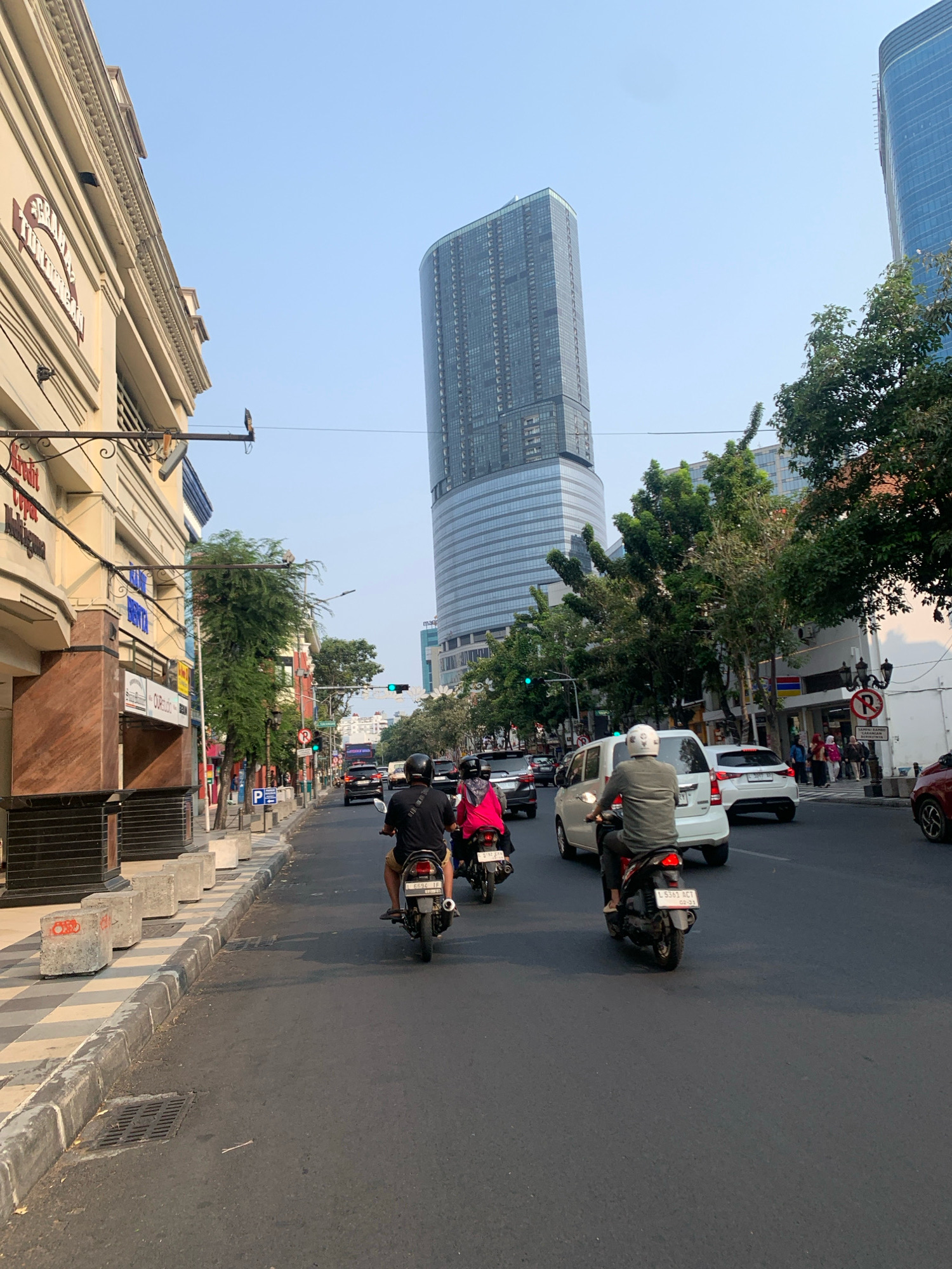 The image shows a street scene in Surabaya, Indonesia, with a tall, modern skyscraper dominating the background. In the foreground, a street is lined with buildings, a sidewalk, and a road with traffic. Several people are riding motorcycles, and cars are visible in the background. The sky is clear and blue, suggesting it is daytime. The buildings on the left appear to be older commercial establishments, while the skyscraper on the right is a contemporary structure. The street is paved and appears to be a busy thoroughfare. There are traffic lights and street signs visible, indicating an urban environment. The overall impression is one of a bustling city street with a mix of old and new architecture.
