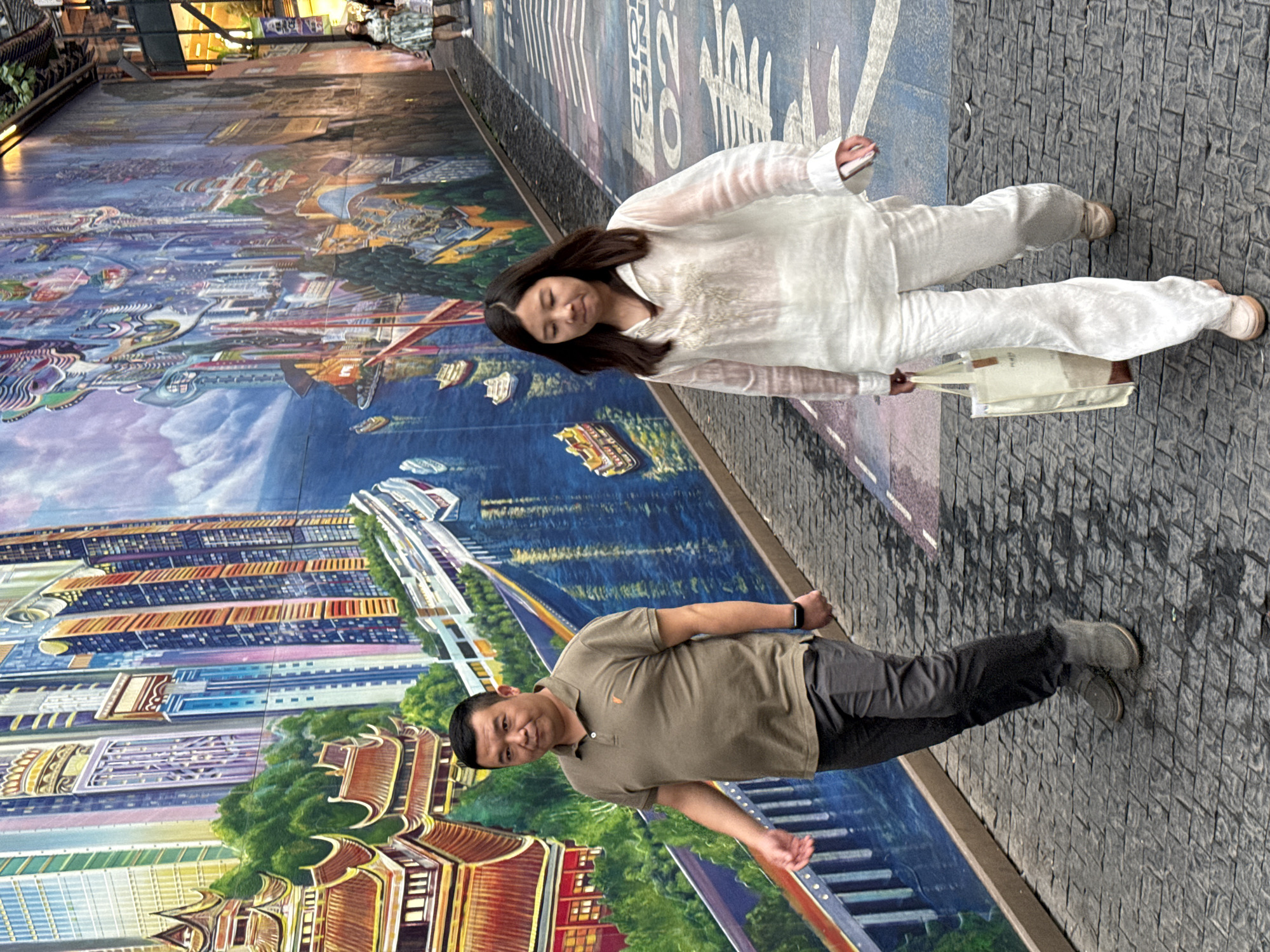 The image shows a man and a woman walking down a street in the Yuzhong District, China, with a vibrant mural covering a building wall on the left. The mural depicts a colorful cityscape with skyscrapers and traditional Chinese architecture, juxtaposed with a river and boats. The man, wearing a brown polo shirt and dark pants, walks slightly ahead of the woman. The woman, dressed in white wide-leg pants and a sheer white blouse, carries a canvas tote bag. The street is paved with dark cobblestones, and there are white painted markings on the ground. In the background, further down the street, another woman in a green dress is visible. The scene appears to be an outdoor urban environment, possibly a pedestrian walkway or alleyway, with the mural serving as a prominent backdrop.