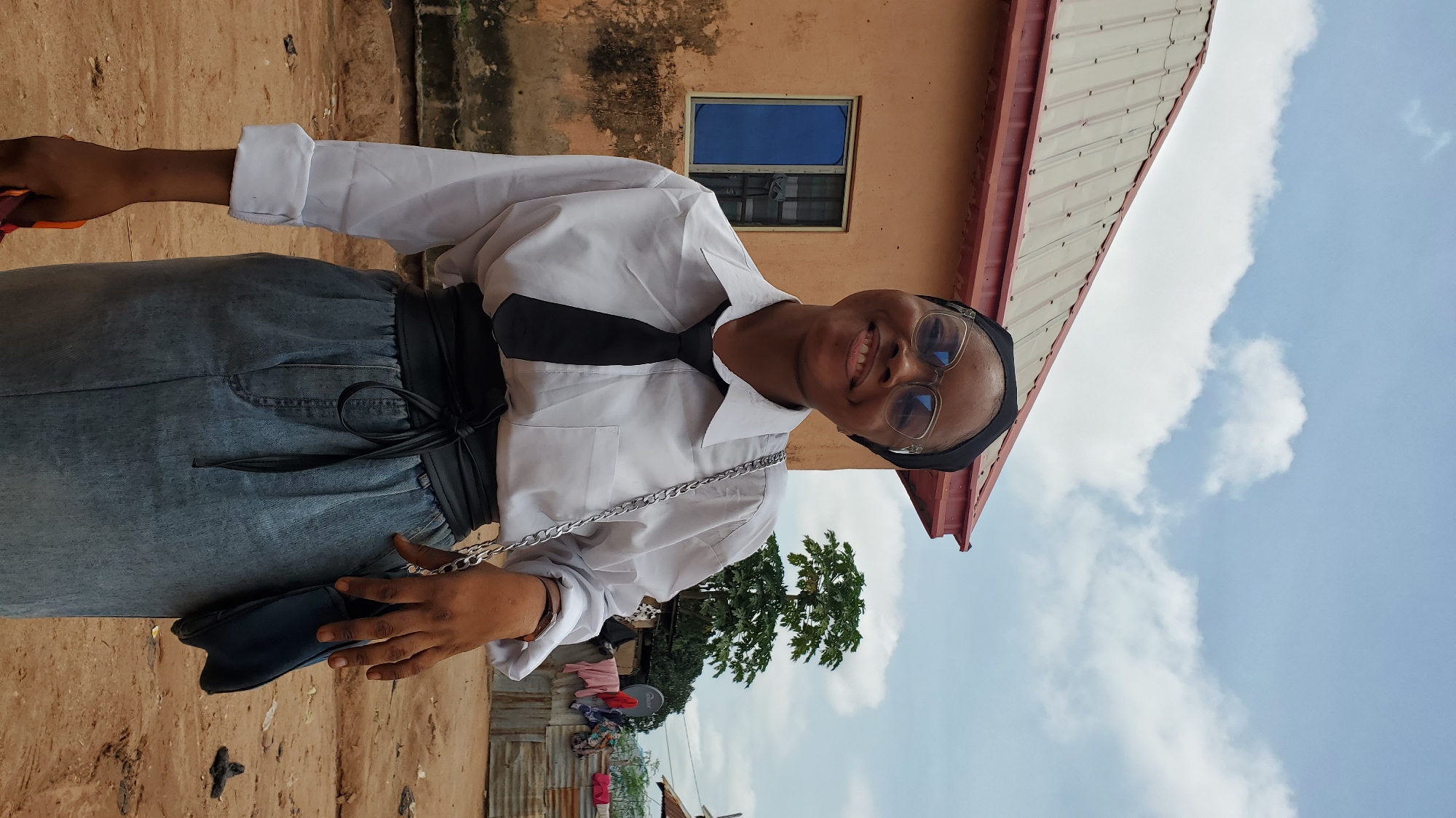 A young woman wearing a black hijab, glasses, a white button-down shirt, a black tie, and a light blue denim skirt stands outdoors in front of a two-story, beige building with a red metal roof in Jalingo, Nigeria. She is smiling and looking towards the camera. The building has a blue-tinted window with a metal grate. The ground is dirt and gravel. In the background, there is a tree, laundry hanging on a clothesline, and what appears to be corrugated metal structures. The sky is partly cloudy.