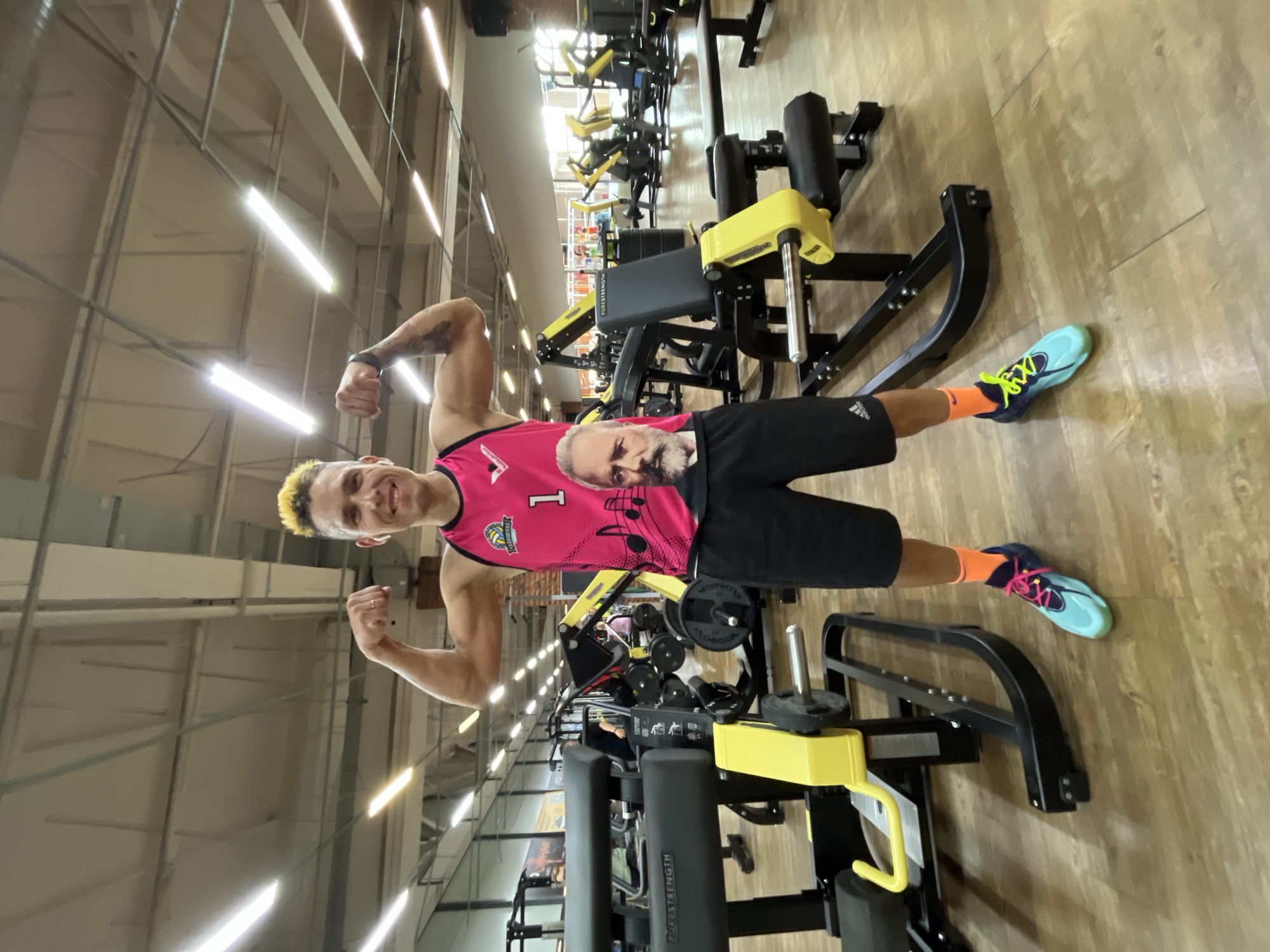 A man is posing in a gym, flexing his biceps. He is wearing a bright pink sleeveless shirt with musical notes and a portrait of a man's face on it, black athletic shorts, bright orange socks, and blue and turquoise athletic shoes. The gym appears to be indoors, with wooden flooring and various weightlifting machines visible in the background. Fluorescent lights illuminate the space from the ceiling. There are other people in the background, but they are out of focus. The location is identified as Chaikovsky, Russia.