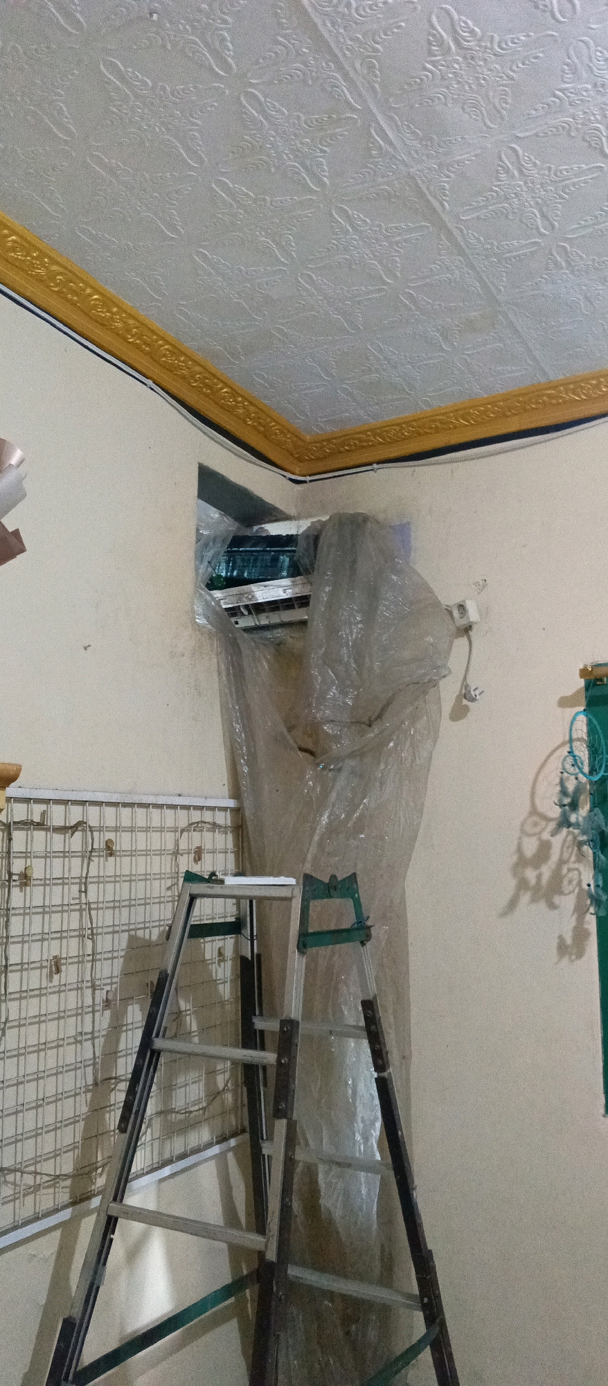 The image captures an indoor scene, likely in Lurah, Indonesia. A portion of a patterned ceiling with a decorative gold trim is visible at the top. The main focus is an air conditioning unit partially covered in clear plastic sheeting, positioned within a wall opening. A silver and green metal ladder stands in front of the air conditioner, suggesting it is either being installed, maintained, or removed. To the left of the ladder is a white metal grid structure with some wires and clips attached. The walls are a light color, and on the right side of the image, a wall outlet and some cables are visible. Further to the right, a green vertical object with shadows of hanging decorations can be seen, possibly a dreamcatcher or similar item. The lighting suggests an interior space, possibly a home or business.