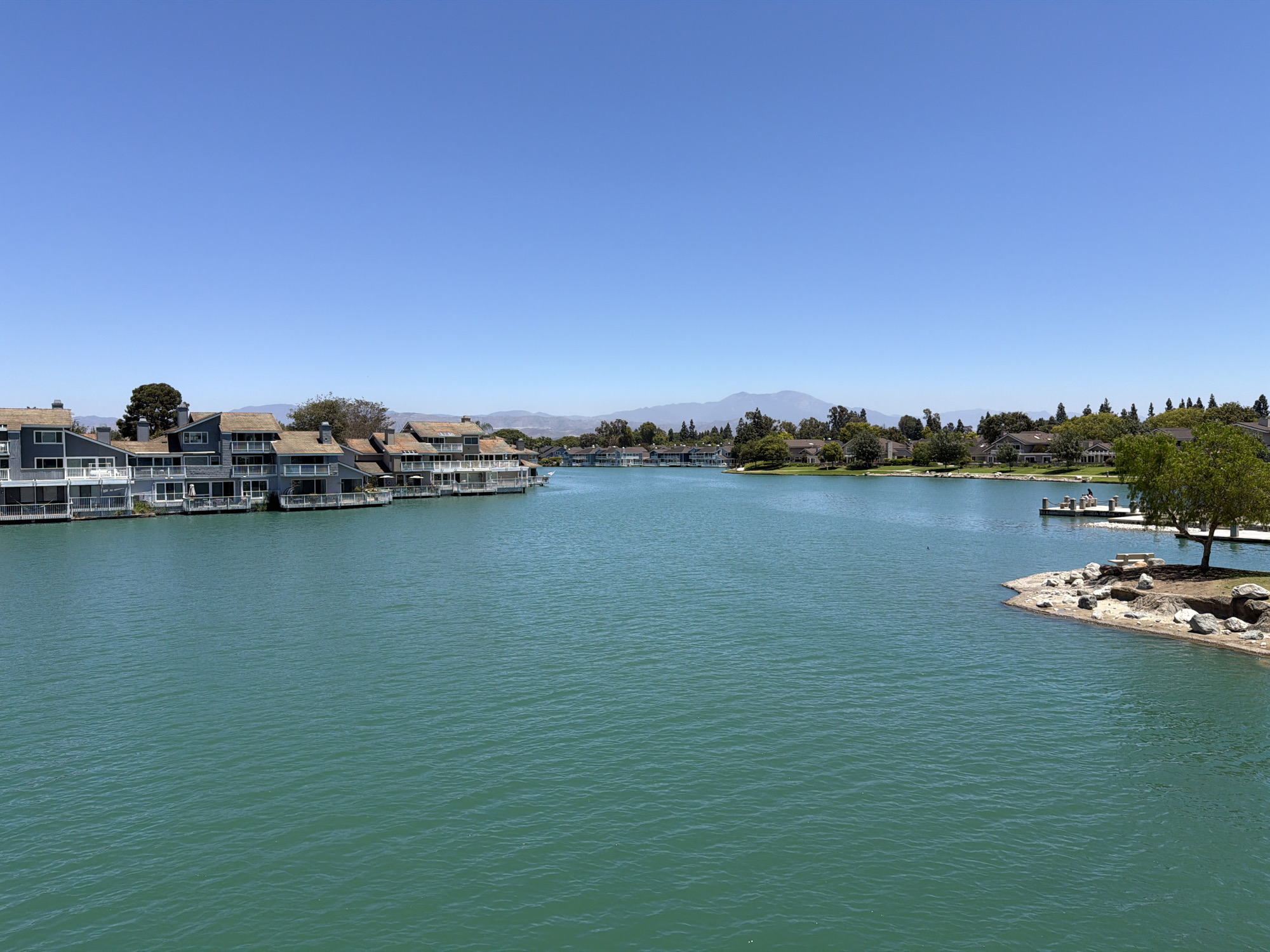This is a wide-angle, eye-level, outdoor, daytime photograph of a man-made lake in the city of Irvine, California, United States of America. The water is a vibrant turquoise color, with ripples indicating a light breeze. On the left side of the frame, a row of multi-story residential buildings with gray siding and brown roofs line the edge of the lake. These buildings feature balconies and large windows facing the water. A small dock with people can be seen on the right side of the lake, next to a tree-lined shore with rocky outcroppings and a park bench. In the background, more residential buildings and trees are visible, with a range of hazy mountains on the horizon under a clear blue sky.
