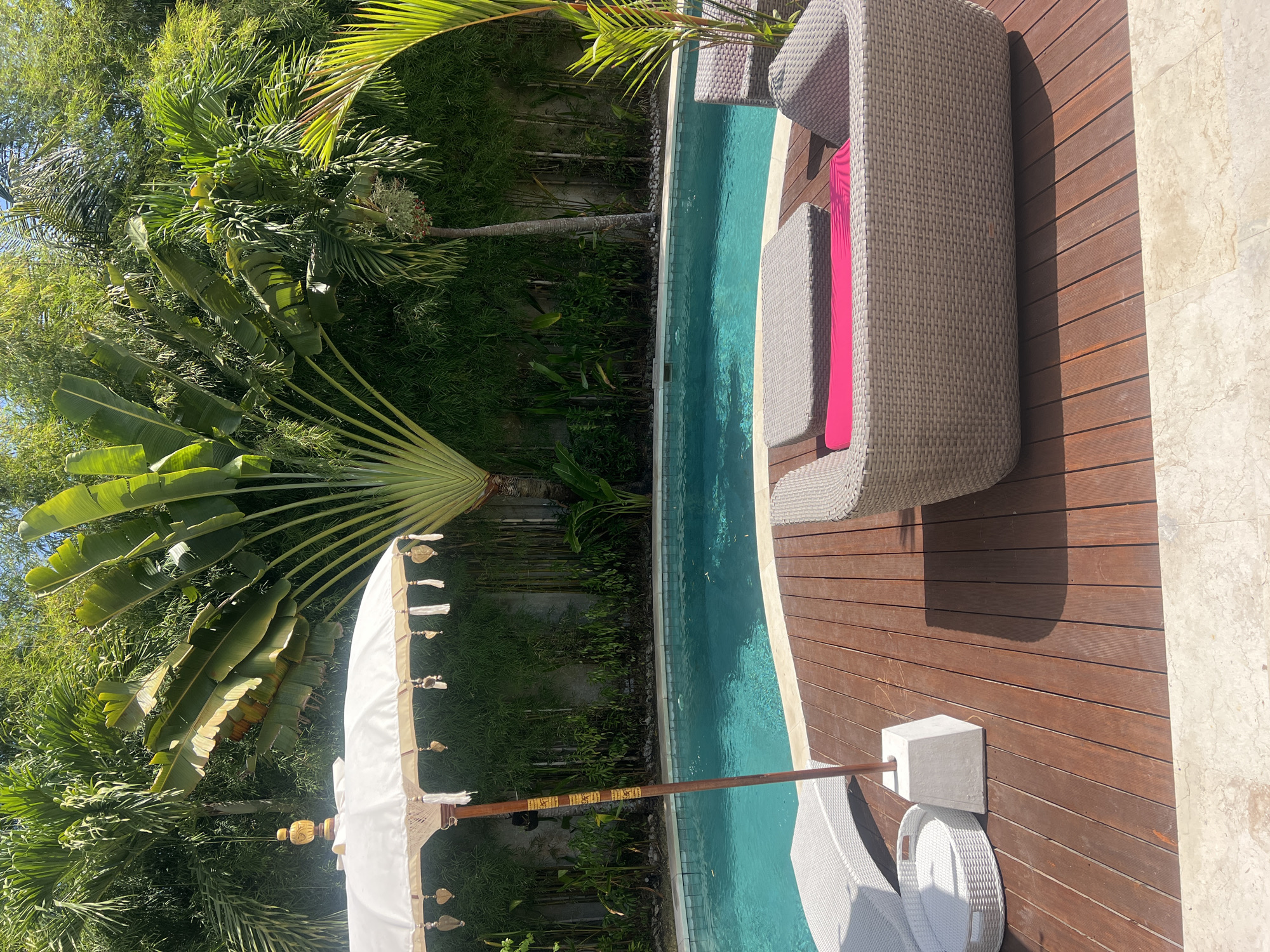 In Kuta Selatan, Indonesia, this image captures a serene outdoor scene with a swimming pool as the focal point. The pool, filled with turquoise water, is bordered by a wooden deck. On the deck, a gray wicker sofa with a bright pink cushion sits invitingly, accompanied by a matching coffee table. To the left, a white wicker tray is placed on the deck near a white sun lounger. A large white umbrella, adorned with decorative tassels and charms, stands prominently, providing shade. Behind the pool, a lush wall of greenery, including bamboo and palm trees, creates a natural and tranquil backdrop. The sunlight casts shadows across the wooden deck, indicating a bright, sunny day.