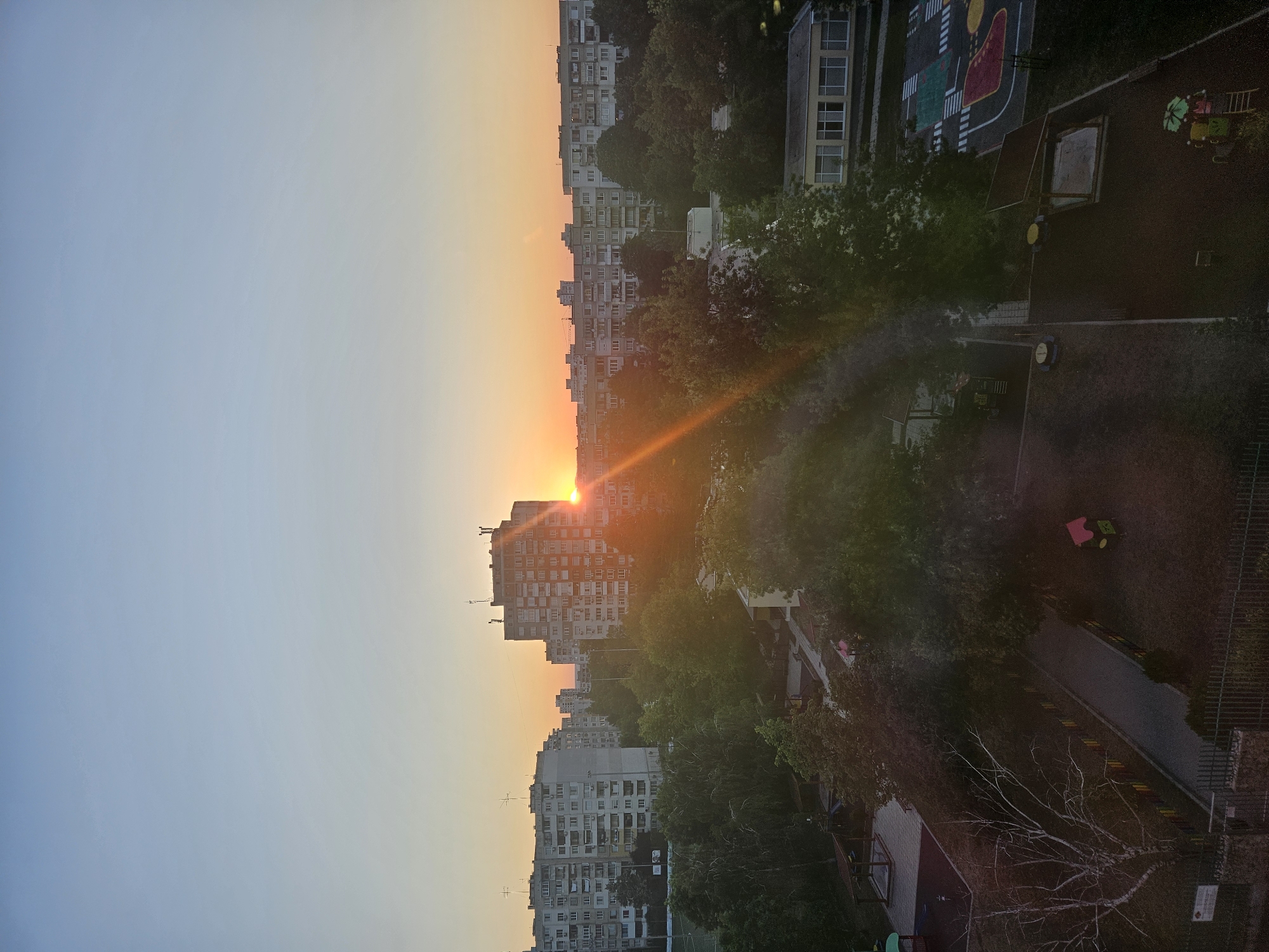 This image depicts a residential area in Sofia, Bulgaria, at sunrise or sunset. The sky is a gradient of light blue to orange, with the sun prominently visible behind a tall, multi-story apartment building. Several other apartment buildings of similar architecture, characteristic of Soviet-era housing blocks, are visible in the background. Dense green trees line the streets and fill the spaces between the buildings. In the foreground, there is a playground with various structures, including a sandbox, slides, and climbing equipment. A fence encloses the area. The scene suggests a typical urban environment with interspersed green spaces.