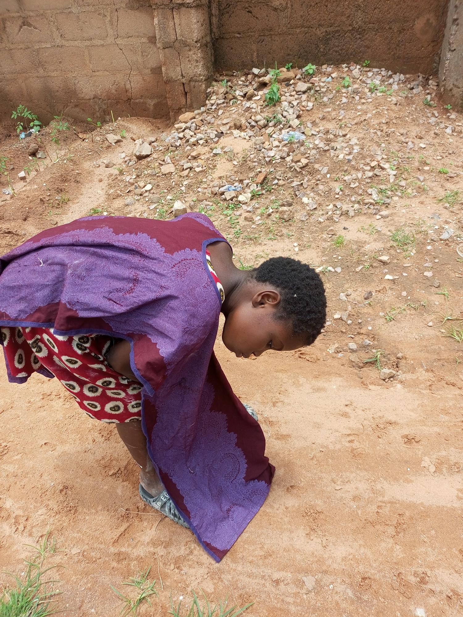 A young Black child, with short, dark, curly hair, is bent over, looking down towards the ground. They are wearing a red and white patterned skirt and a large, purple, patterned wrap that drapes over their shoulders and body. The child's feet are visible, wearing dark sandals. The child is positioned on sandy ground, which is interspersed with small stones and sparse green vegetation. Behind the child, a roughly constructed wall made of large, rectangular bricks forms the background. The wall appears weathered and cracked. To the right of the wall, there is a pile of stones and dirt. The overall environment suggests an outdoor setting, possibly in a more rural or undeveloped area of Tempure, Nigeria.